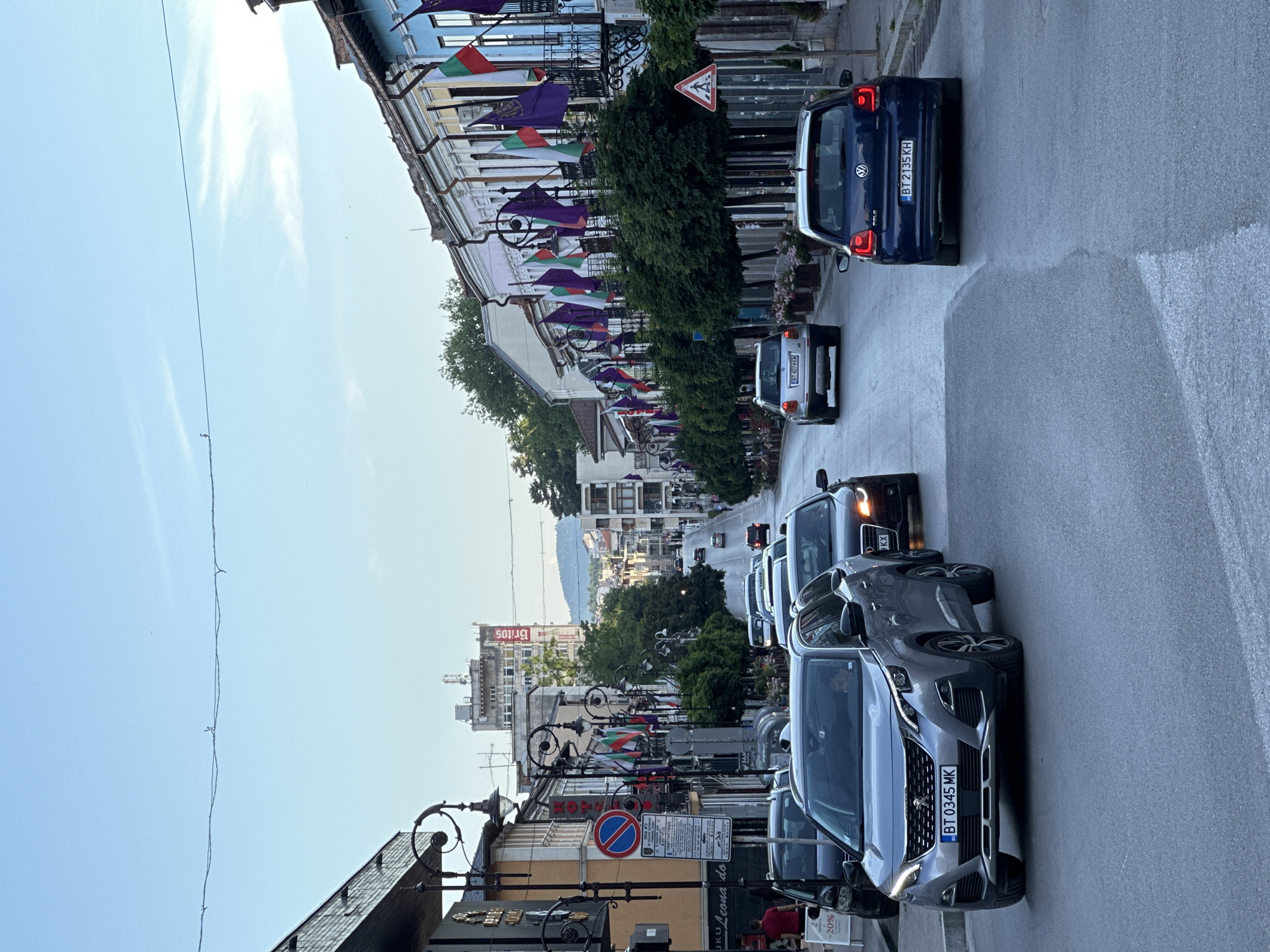 The image depicts a street scene in Veliko Tarnovo, Bulgaria, on a cloudy day. The street is lined with buildings displaying a row of colorful flags, alternating between green, white, and red, and purple flags, possibly indicating a local celebration or event. The buildings have balconies with ornate railings. Trees are planted along the sidewalk, some of which are shaped into spheres.
Traffic is present on the street, with several cars visible. In the foreground, a silver Peugeot SUV is stopped in traffic, with its license plate reading "BT 0345 MK". To its right, a dark Audi sedan is also stopped. Further down the street, other vehicles are moving, including a dark blue Volkswagen Polo with license plate "BT 2135 KH".
A pedestrian crossing sign is visible on the right side of the street. In the background, a tall, light-colored building with the word "BRITOS" is prominent. The street appears to be a main thoroughfare with shops and businesses lining its sides. The overall impression is a bustling urban environment with a festive atmosphere due to the flags.