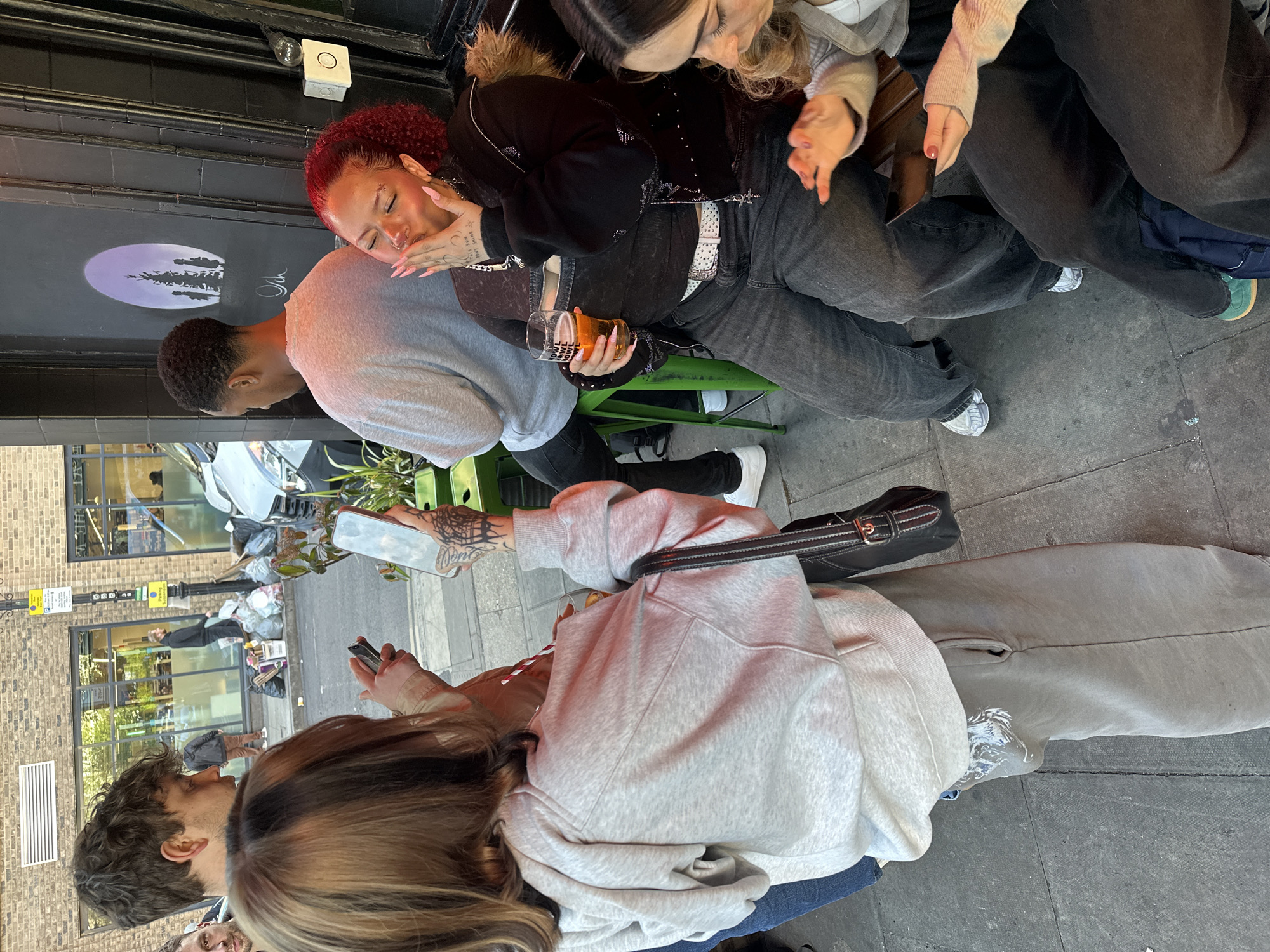 The image shows a group of people outside a building in London, United Kingdom. A woman with red curly hair and long nails is holding a pint glass, resting her face on the shoulder of a man in a grey sweatshirt. Other people are seated nearby. A woman with tattoos on her arm is holding a mobile phone. The building in the background has a dark grey facade with a sign featuring a moon and trees. The street scene includes reflections in the glass of a building across the street, showing pedestrians and refuse bags.