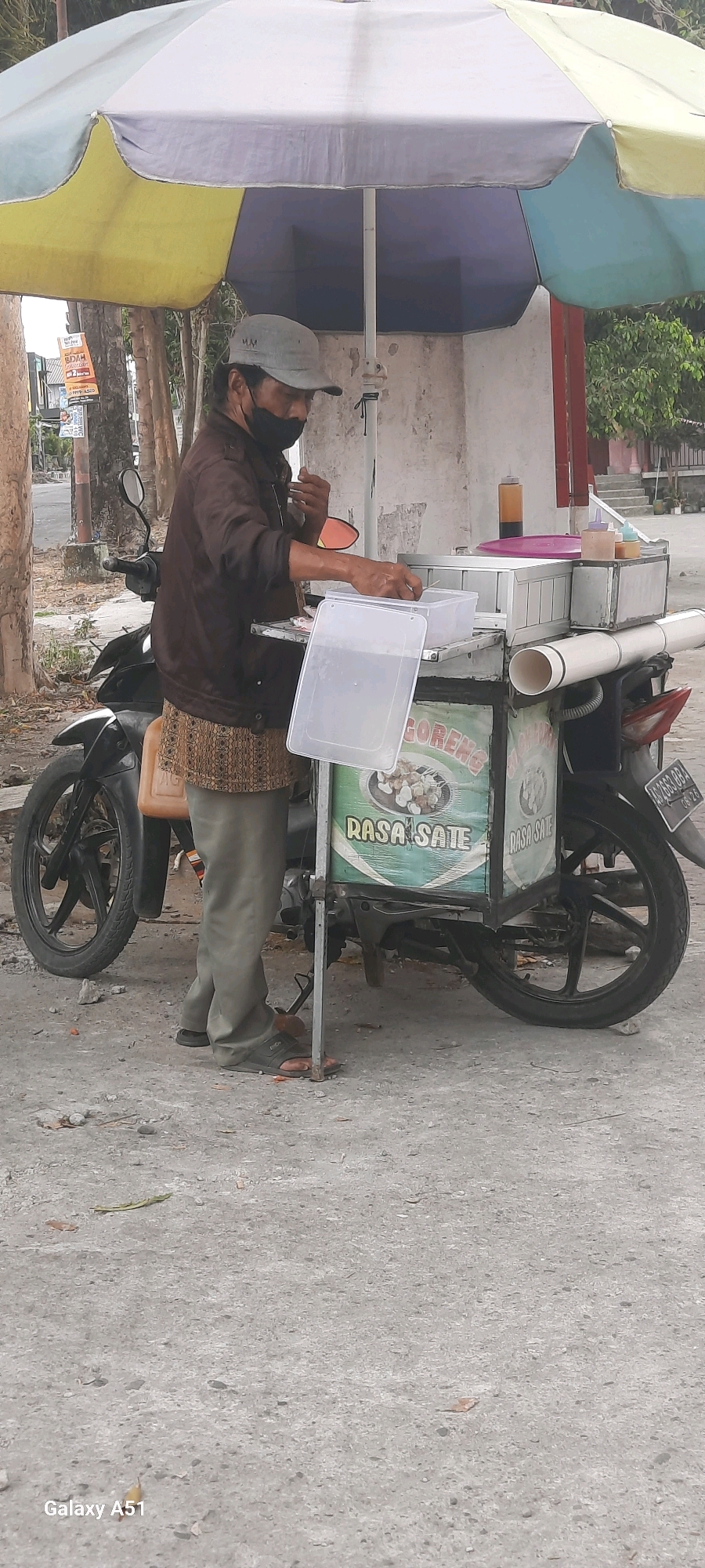In the city of Tangkil, Indonesia, a street vendor, wearing a gray cap, a dark brown jacket, and light green pants, is preparing food at a mobile cart. The cart is attached to a black motorcycle and covered by a large umbrella with yellow, blue, and white sections. The vendor is holding tongs and appears to be serving food from a container. The cart has a sign that reads "GORENG RASA SATE". There are various bottles and containers on the cart's top surface. The ground is paved with concrete and scattered with debris. The overall scene depicts a typical street food vending operation.