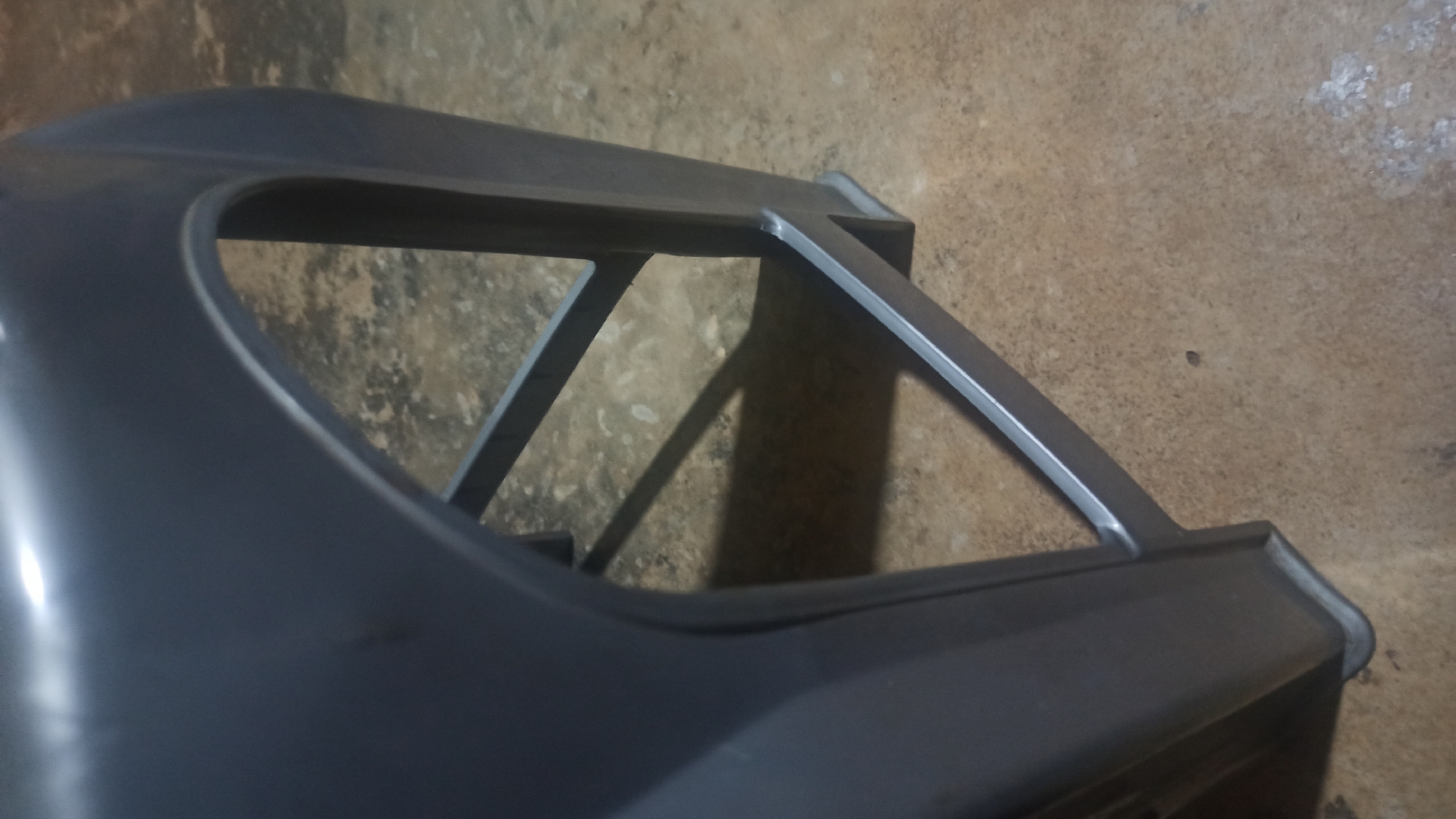 A close-up, low-angle shot captures the gray plastic leg and frame of a stool, situated indoors on a textured, light brown floor. The stool's leg is rounded at the top and rectangular at the base, with support beams forming an arched opening. A shadow is cast on the floor, indicating a light source from the upper left. The overall environment appears to be an indoor space in Tuba, Indonesia, though specific details about the city are not visible. There are no people or discernable actions depicted in the image.
