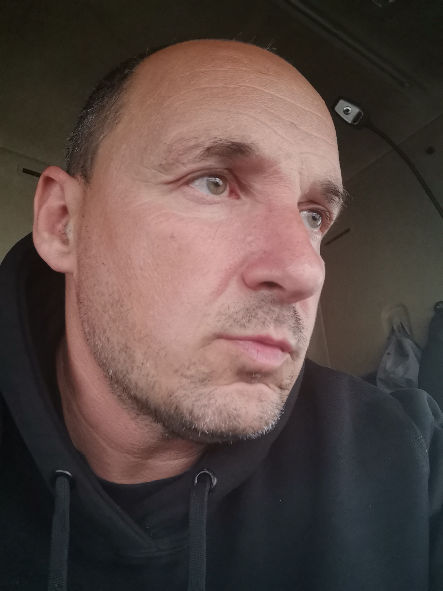 The image presents a close-up, head-and-shoulders portrait of a man, likely in his middle age, captured from a slightly low angle. He has fair skin, a receding hairline with short, dark, and subtly graying hair on the sides. A light stubble covers his chin, jawline, and upper lip. His eyes, light in color (possibly hazel or green), are wide and focused intently towards the viewer's right, giving him a serious, perhaps pensive or observant expression. He is wearing a dark, presumably black, hooded sweatshirt, with the drawstrings visible around the neckline.
The background is an interior space, appearing dim and muted in tone, consistent with a vehicle cabin. In the upper right corner, a small, dark grey electronic device with a metallic-like, rectangular surface and a central circular element is visible. This device, possibly a light or camera, is connected by a dark wire extending upwards, suggesting it's mounted to an overhead surface. In the lower right background, some crumpled, light-colored fabric or clothing is partially discernible. The overall lighting is subdued, indicating an indoor setting or low-light conditions. No visible text is present in the image. The man is likely in or around Karapelit, Bulgaria, possibly within a vehicle cabin, engaged in observation or thought.