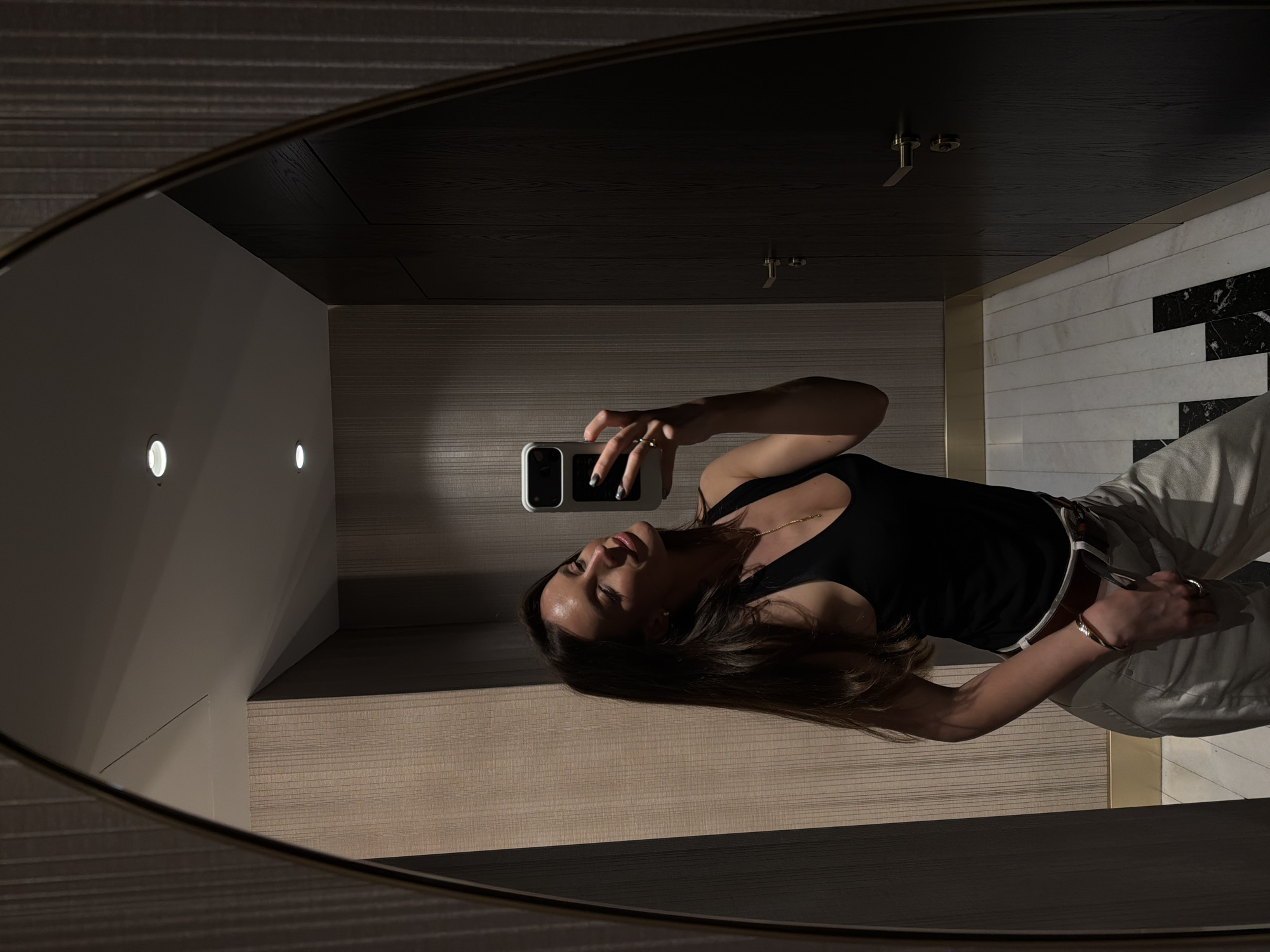 A woman takes a selfie in a mirror with a brass frame in a modern bathroom. The walls are decorated with light brown striped tiles on one side and white tiles with black marble accents on the other. The ceiling is dark brown with recessed lighting and sprinkler heads. The woman wears a black tank top and light-colored pants with a belt. The location is likely in Budva, Montenegro, given the architectural style.
