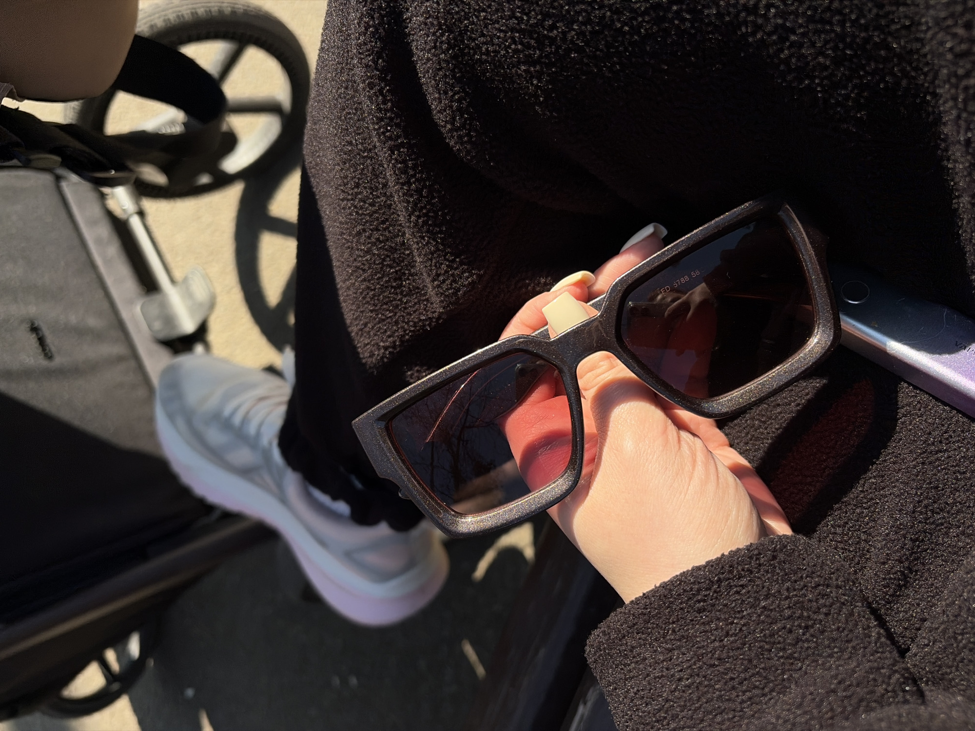 A photograph taken in Krasnodar, Russia, features a person holding a pair of dark-framed sunglasses in their right hand. The sunglasses have rectangular lenses with a dark tint. The person is wearing a black textured fabric, possibly a jacket or coat. In the background, a stroller with grey fabric is visible, and part of a light-colored sneaker with a pink sole is seen next to it. The setting appears to be outdoors, likely on a sunny day, given the shadows and lighting. A metallic object, possibly a phone, rests next to the sunglasses. The hand holding the glasses has manicured nails, painted with a light yellow color.