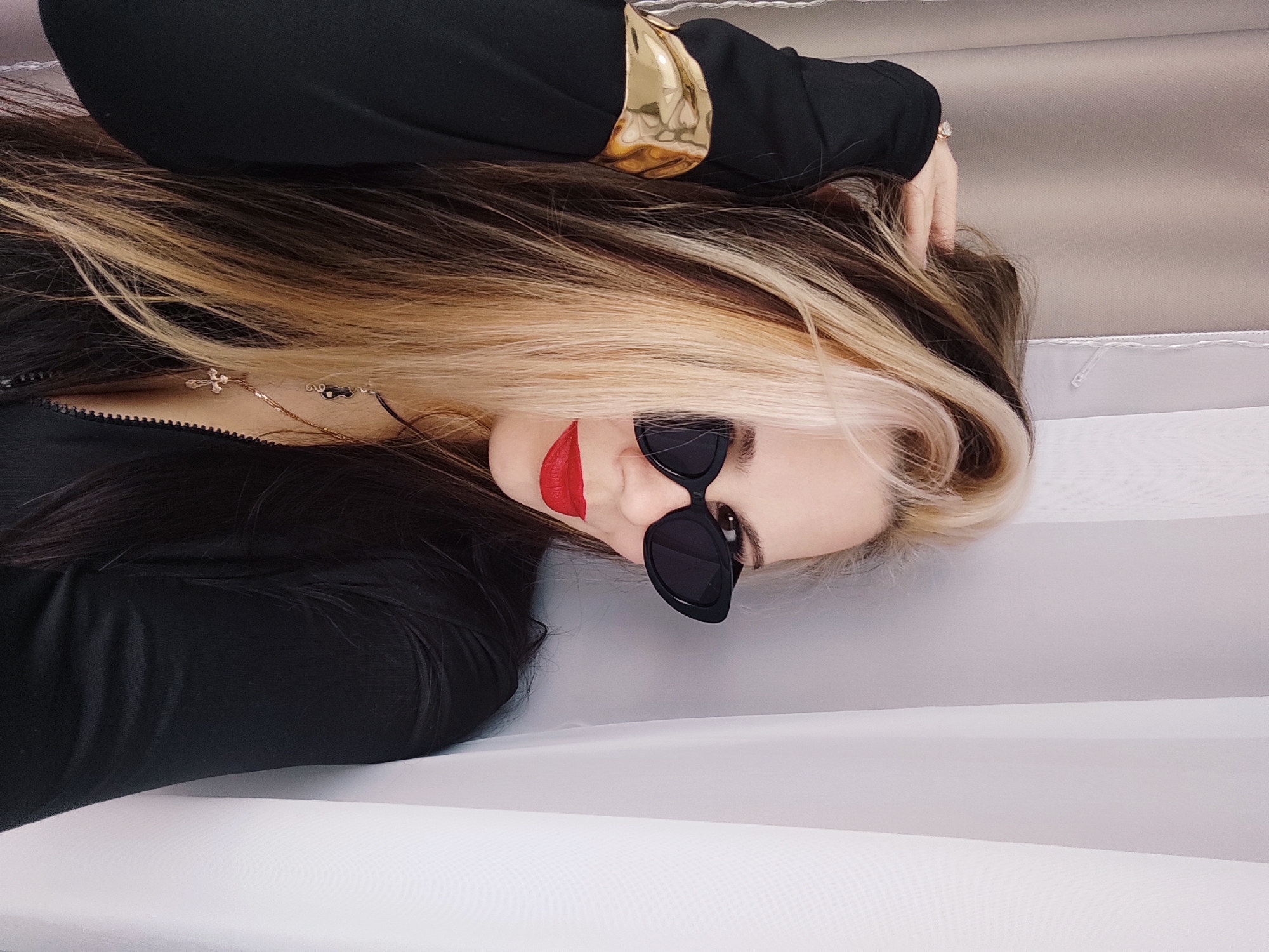 A woman with black and blonde hair, wearing sunglasses and red lipstick, is lying on a white surface. She is dressed in a black top with a gold accent on the sleeve. She has a necklace on. The location is Chaikovsky, Russia.