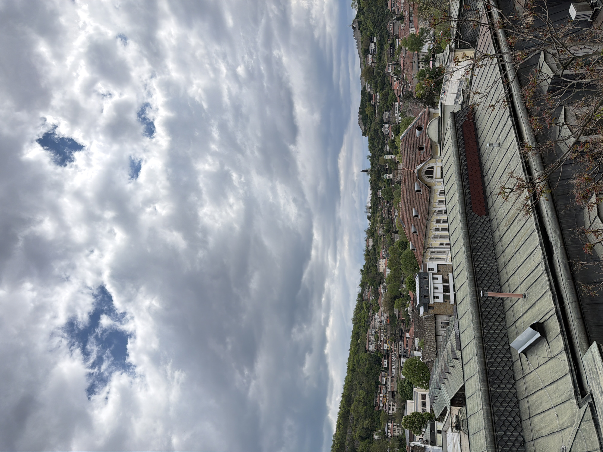 The image shows a cityscape from Lovech, Bulgaria, taken from an elevated position, possibly a rooftop. In the foreground, a complex of buildings with grey-green metal roofs and red-tiled sections extends into the frame. A bare tree branch with sprouting buds is visible on the right.
Beyond the rooftop buildings, a densely populated hillside slopes downward. Houses with a mix of red-tiled and other roof types are interspersed with trees. In the background, a ridge topped with some sort of fortifications or ruins is visible.
The sky dominates a large portion of the image, filled with a thick layer of mostly grey clouds, with patches of blue sky visible in the lower left corner. The overall lighting suggests an overcast day.
