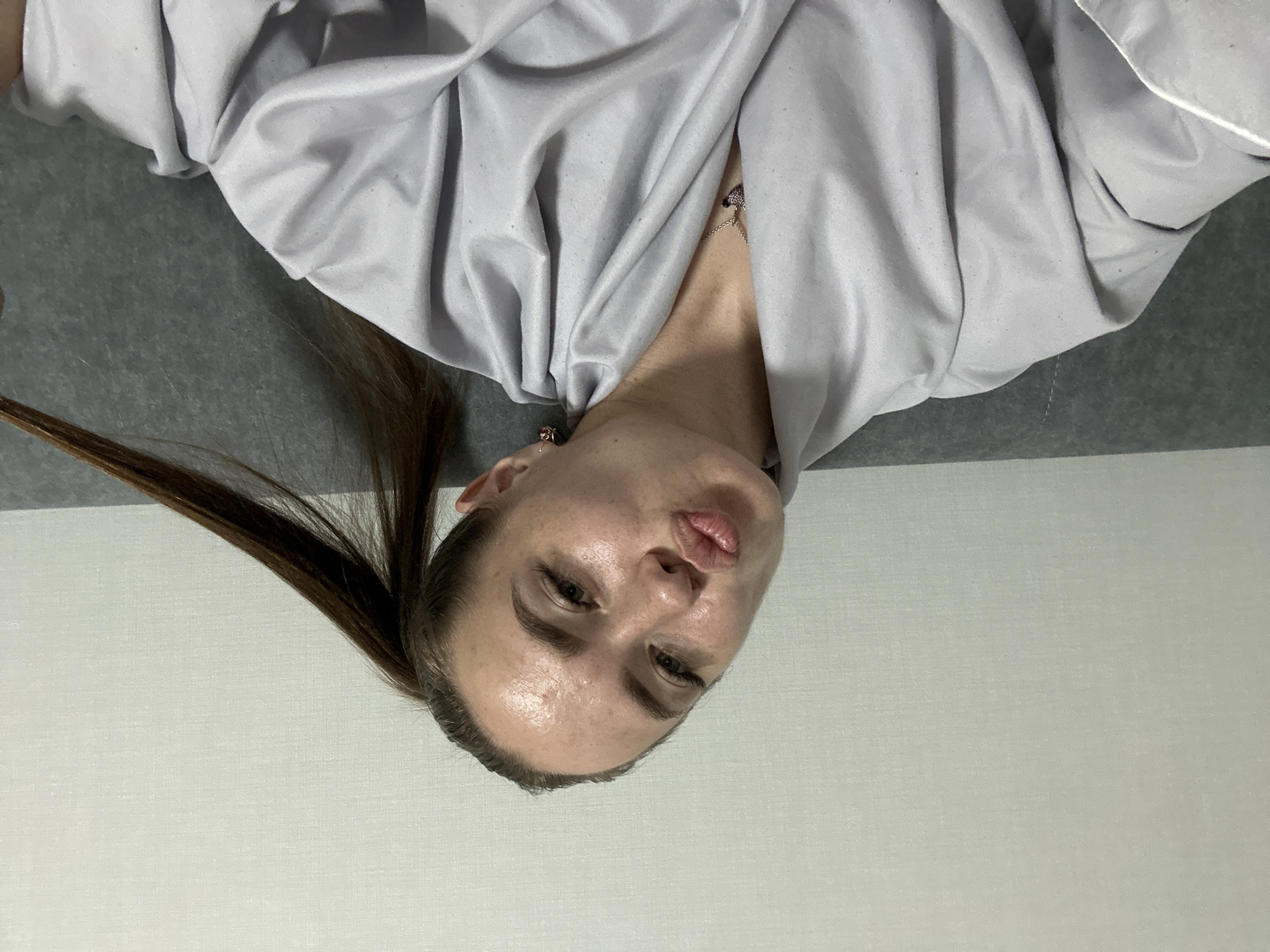 A woman is shown from the chest up, wearing a gray robe. Her hair is pulled back into a ponytail, and she is holding a strand of her hair. She is looking towards the camera with her lips pursed. The background is a plain, textured wall. The scene appears to be indoors, possibly a bedroom or living room.