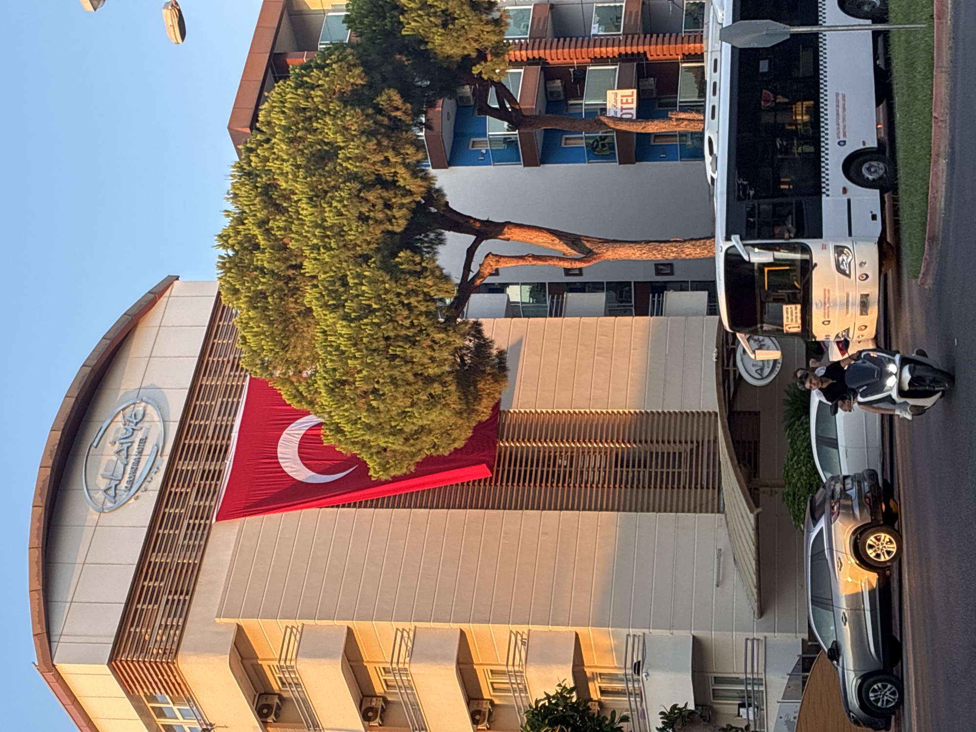 An outdoor, eye-level, medium shot captures a street scene in Alanya, Turkey, at dusk. A large building with the "ALANYA CLEOPATRA HOTEL" sign is prominently featured, displaying a Turkish flag hanging vertically down its facade. To the right, a modern multi-story building with blue balconies and a sign reading "HOTEL" is visible. Two large evergreen trees partially obscure the buildings. In the foreground, a white bus is stopped at a traffic light, and next to it, a person on a scooter rides past. A silver SUV and a white car are parked on the street. The lighting suggests the golden hour, with warm sunlight illuminating the buildings.