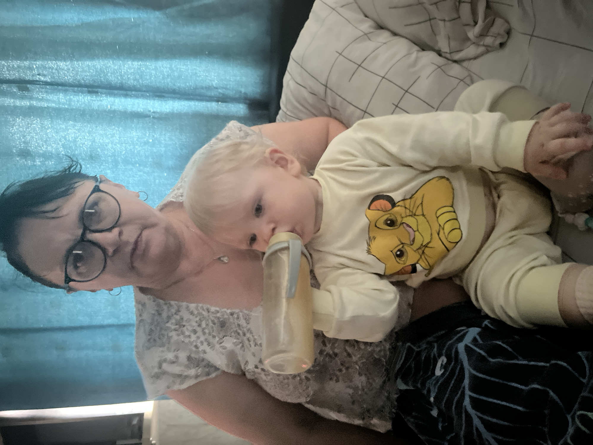 The media file shows a woman and a baby indoors. The woman is wearing glasses and a lacy top, while the baby is dressed in a light yellow onesie with a graphic of Simba from The Lion King. The baby is holding and drinking from a bottle, resting on the woman's chest. The setting appears to be a bedroom or living room, with a blue curtain visible in the background and a pillow with a grid pattern. The lighting suggests it could be daytime, indoors. The scene conveys a sense of care and comfort between the woman and the baby. There is no visible text in the image. The location context is given as Фоки, Russia.