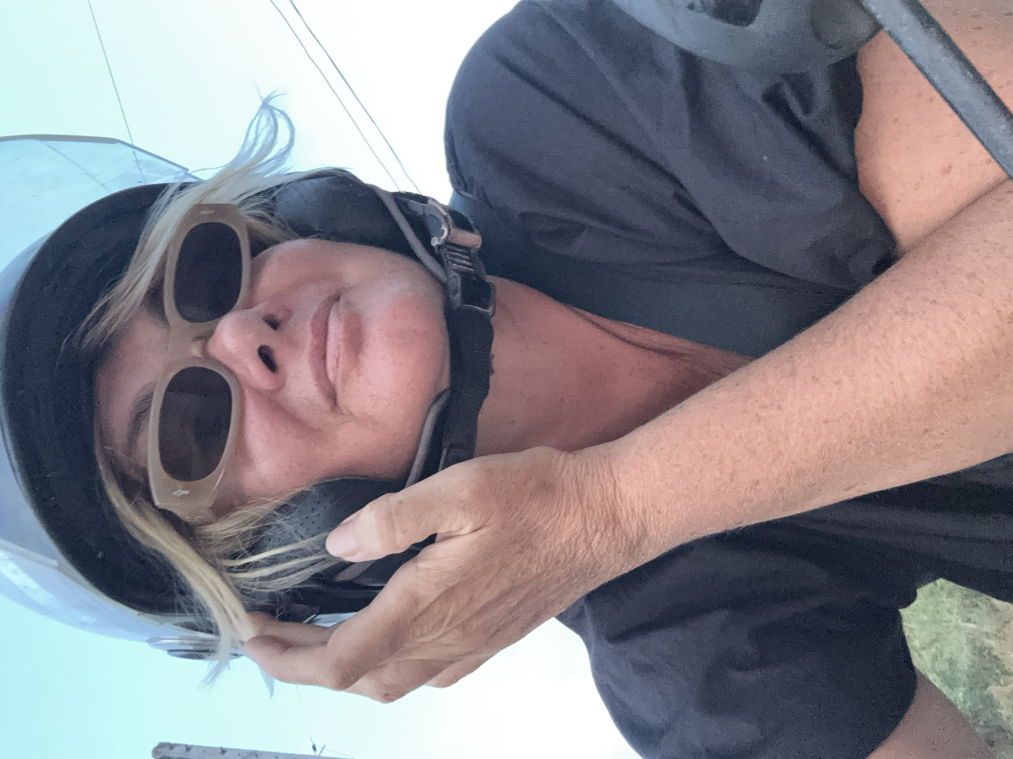 A person wearing a helmet and sunglasses is shown in a close-up shot. The person's face is the main focus, with a hand near the helmet's ear cover. A black shirt is visible. The background is out of focus, showing what appears to be an open sky. The location in the city of บ้านบางรักษ์, Thailand, cannot be verified from the image.