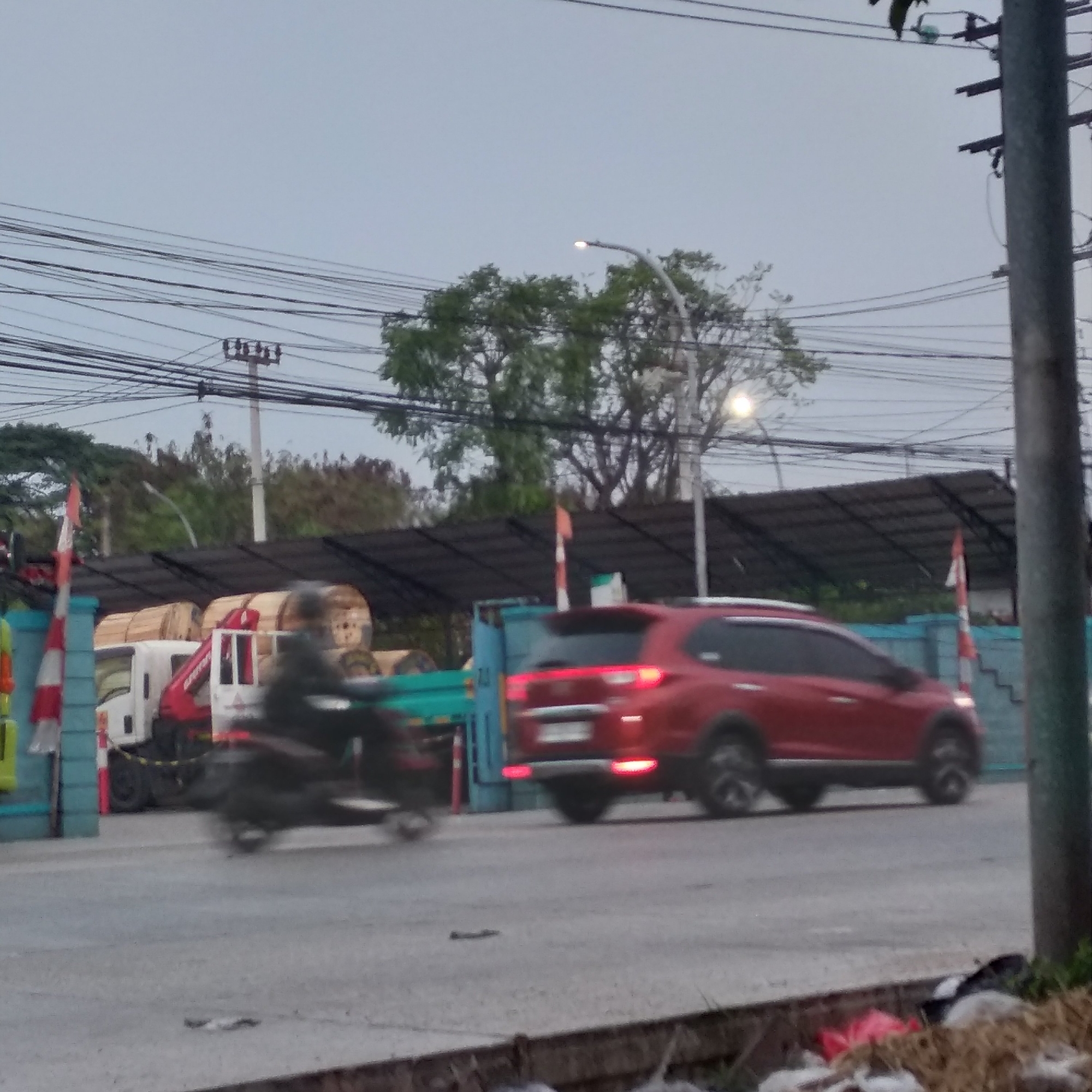 The image captures a blurred street scene in Balaraja, Indonesia, under an overcast sky. In the foreground, a red SUV is in motion, its taillights illuminated. To the left, a motorcycle with a rider is also moving, appearing as a dark blur. Behind the vehicles, a white truck with a red crane arm is parked, and large wooden spools are stacked. A blue fence and a red and white striped pole are visible on the left side of the frame. Overhead, a complex network of electrical wires crisscrosses the sky, supported by utility poles. Trees with dense green foliage are scattered across the background, and a street lamp with a glowing bulb is positioned above the trees. The ground is paved and appears somewhat worn, with some debris visible along the curb in the bottom right corner of the image.