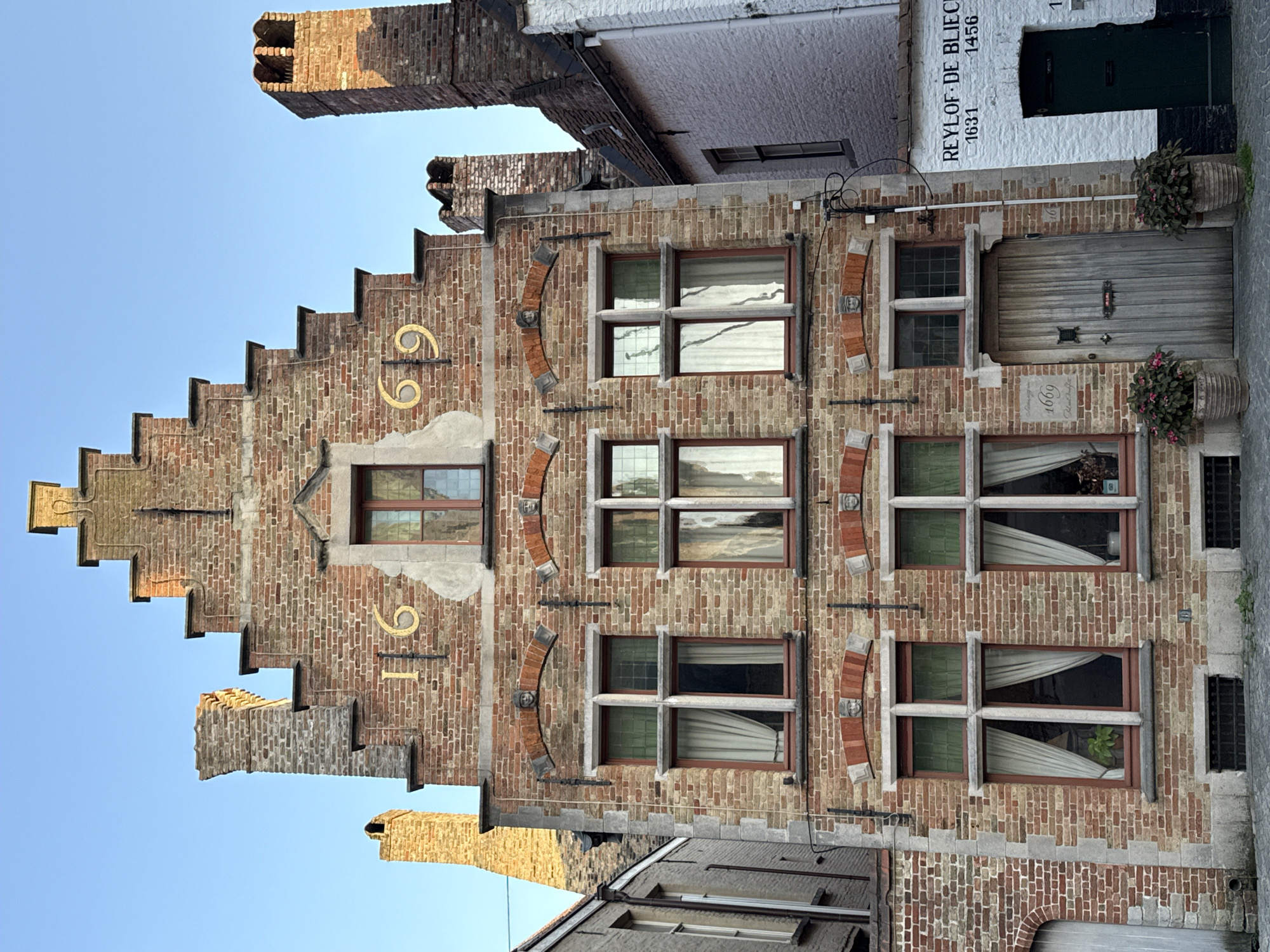 This is a vertical, close-up shot of the ornate brick facade of a building in Bruges, Belgium. The building features a stepped gable roofline and multiple windows with decorative brickwork. The facade is constructed from reddish-brown bricks, with lighter stone accents around the windows and the stepped gable.
Golden numbers "69" are prominently displayed on the left side of the building, suggesting a house number or a historical marker. Below the "69", the number "91" is also visible in gold. The windows are multi-paned and have dark wooden frames. Some windows have white curtains, and one window on the right shows a potted plant.
The building to the right is painted white and has dark green doors. Text is visible on the white building, reading "REYL OF DE BLIECK" followed by numbers "1631-1456". Another number "1669" is visible near the ground floor entrance on the brick building.
The scene appears to be a street in Bruges, likely a historical area, given the architecture. The lighting suggests it is daytime, possibly late afternoon due to the warm light illuminating the brickwork and casting shadows. The sky visible in the upper left corner is clear and blue, indicating fair weather. There are no people visible in the frame. The overall impression is one of historical charm and architectural detail.