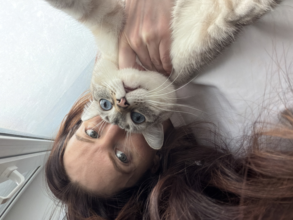 A close-up, angled portrait features a female subject and a domestic cat in an indoor environment. The upper right portion of the female's face is visible on the left, characterized by a light green eye directed towards the camera and long, wavy, dark brown hair. She is wearing a white top. The cat, positioned centrally, is a light-colored domestic shorthair with blue eyes and prominent white whiskers, its head tilted upwards, also looking directly into the camera with a slightly open mouth. Human hands are visible holding the cat around its torso. The background is a brightly lit, out-of-focus area, consistent with a window showing an overcast sky or bright exterior, with a portion of a white window frame visible at the top. The specific location within Perm, Russia, is not discernable from the visual content of the image.
