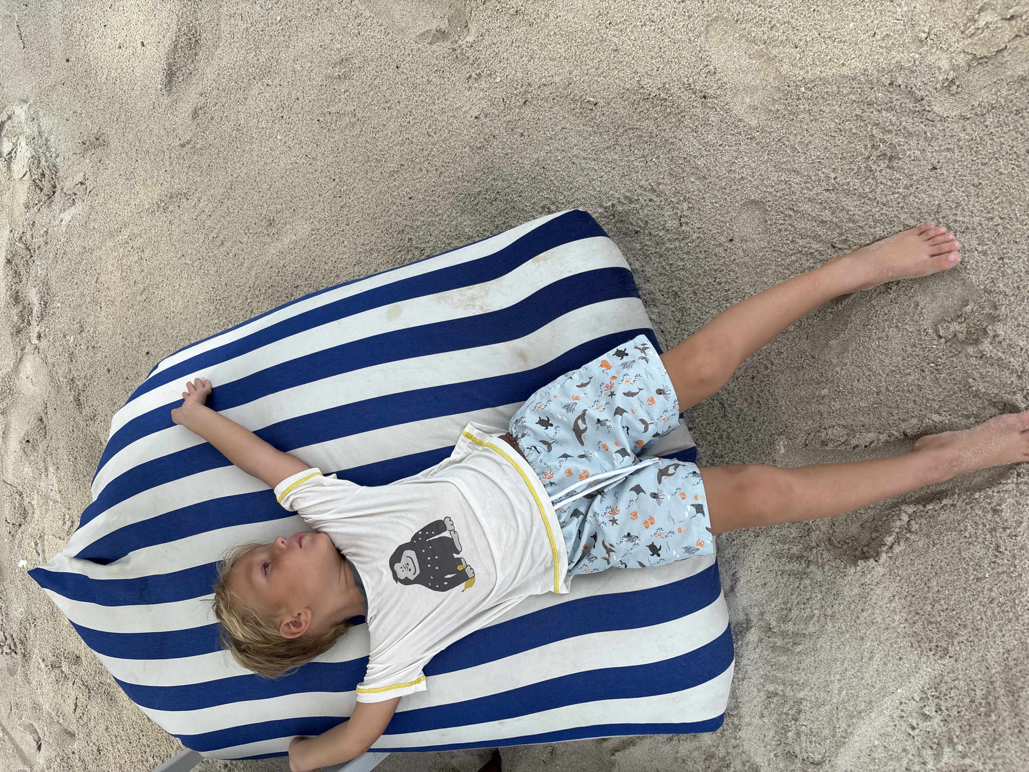 A child lies on a blue and white striped cushion on a sandy surface. The child has blonde hair and is wearing a white shirt with a gorilla graphic and blue shorts with a sea life pattern. The child's arms are outstretched, and their legs extend onto the sand. The location is Baan Chaweng, Thailand.