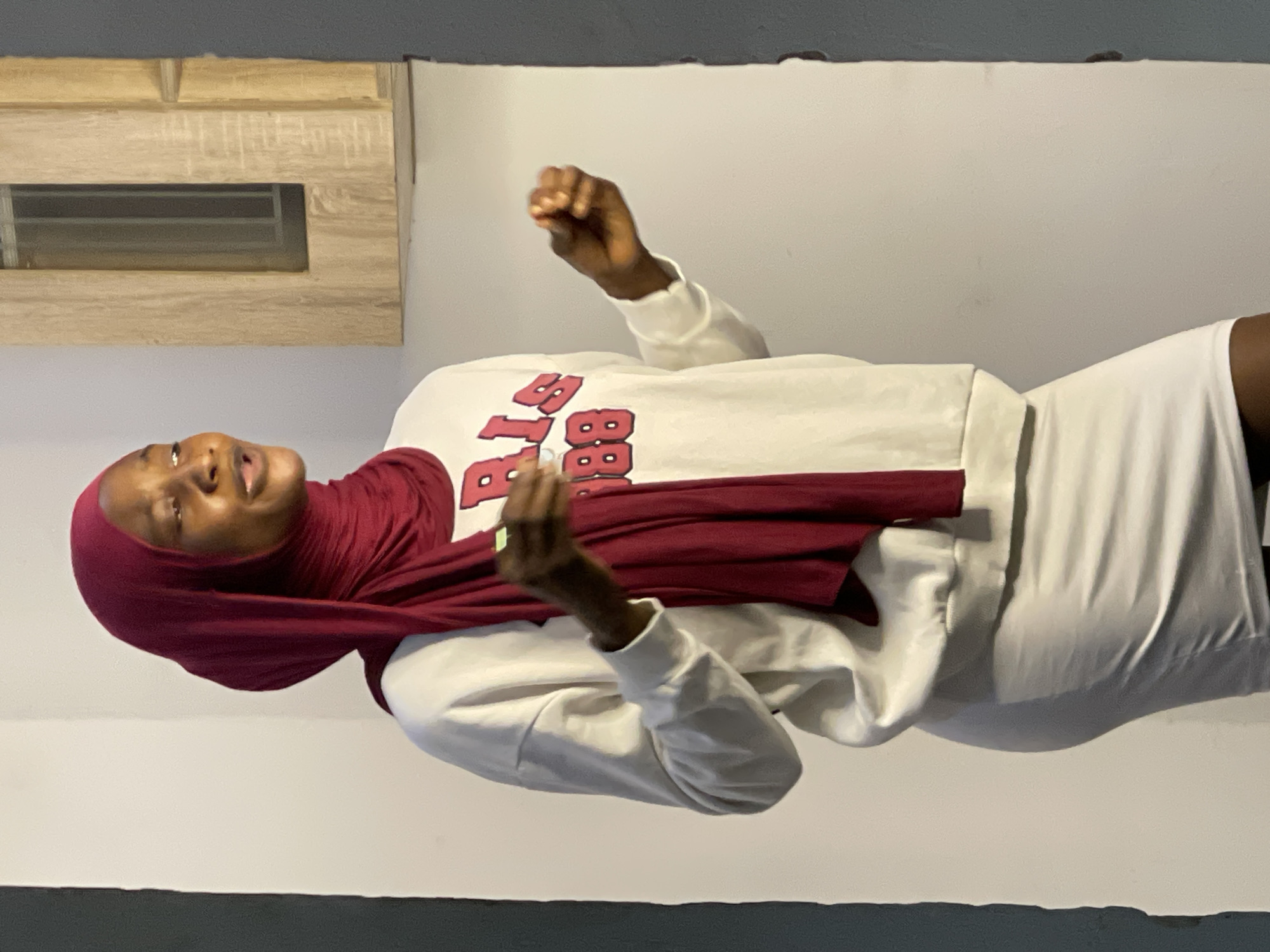 A young woman wearing a maroon hijab and a white sweatshirt with red lettering that reads "PARIS 1888" and a white skirt stands in an indoor setting. She is looking to her right with her mouth open, as if speaking or singing, and has her hands raised and slightly bent. A wooden cabinet with a dark rectangular window is visible on the wall behind her. The wall is painted a light color, and a dark gray column is on the right edge of the image. The lighting appears to be artificial, creating shadows on the wall. The scene is likely in a residential or commercial space within the city of Abuja, Nigeria, given the context provided.