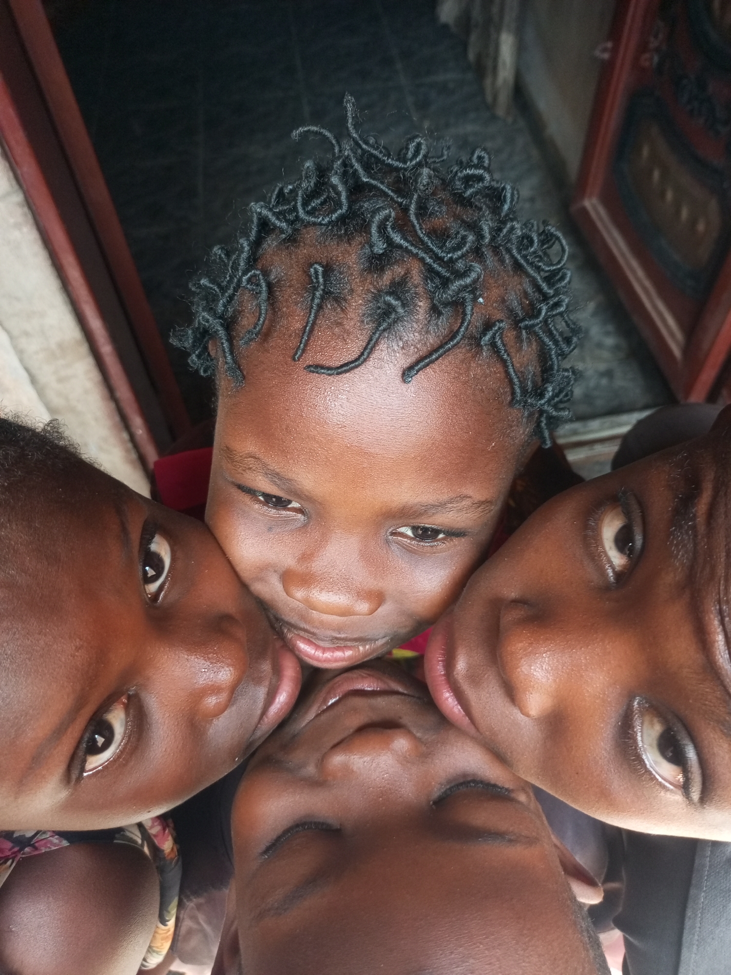 The image is a close-up, high-angle shot of four young Black individuals pressed together, their faces almost touching. Their skin is a rich brown, and their hair is styled in small twists. The person in the center is looking directly at the camera with a slight smile. The other three individuals are angled in various directions, with their eyes closed or looking away. The setting appears to be indoors, with a dark doorway and a wooden frame visible in the background. The floor is tiled. There is no specific indication of the exact location within Gombe, Nigeria, beyond it being an urban or semi-urban environment.