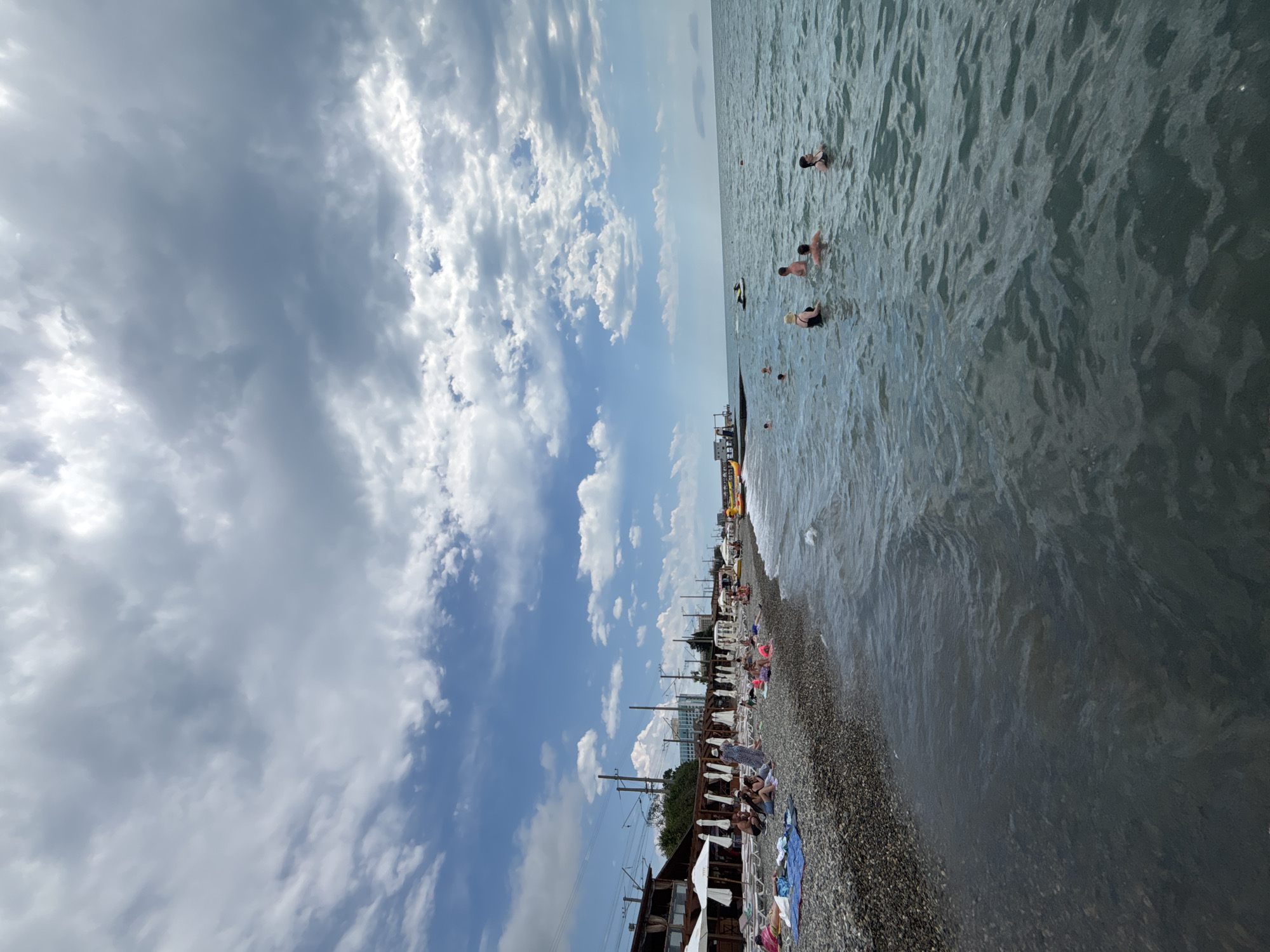 The image depicts a bustling beach scene in Sochi, Russia, under a partly cloudy sky. The sun is likely high in the sky, indicated by the bright daylight. The sea is a choppy, dark blue, with several people swimming and wading in the water. Some are further out, enjoying the open water, while others are closer to the shore. A jet ski is visible in the water, suggesting recreational activities.
The beach itself is covered in pebbles and lined with numerous beach umbrellas and lounge chairs, many of which are occupied by sunbathers. People are scattered across the beach, either relaxing on towels or engaging in conversations. In the background, there are some buildings and structures, possibly hotels or beachside establishments, and electrical wires suggesting nearby infrastructure. The overall atmosphere appears to be one of leisure and enjoyment, typical of a summer day at the seaside. The clouds are a mix of fluffy white and darker grey patches, hinting at the possibility of changing weather, but the general lighting suggests a pleasant day. No visible text is present in the image.