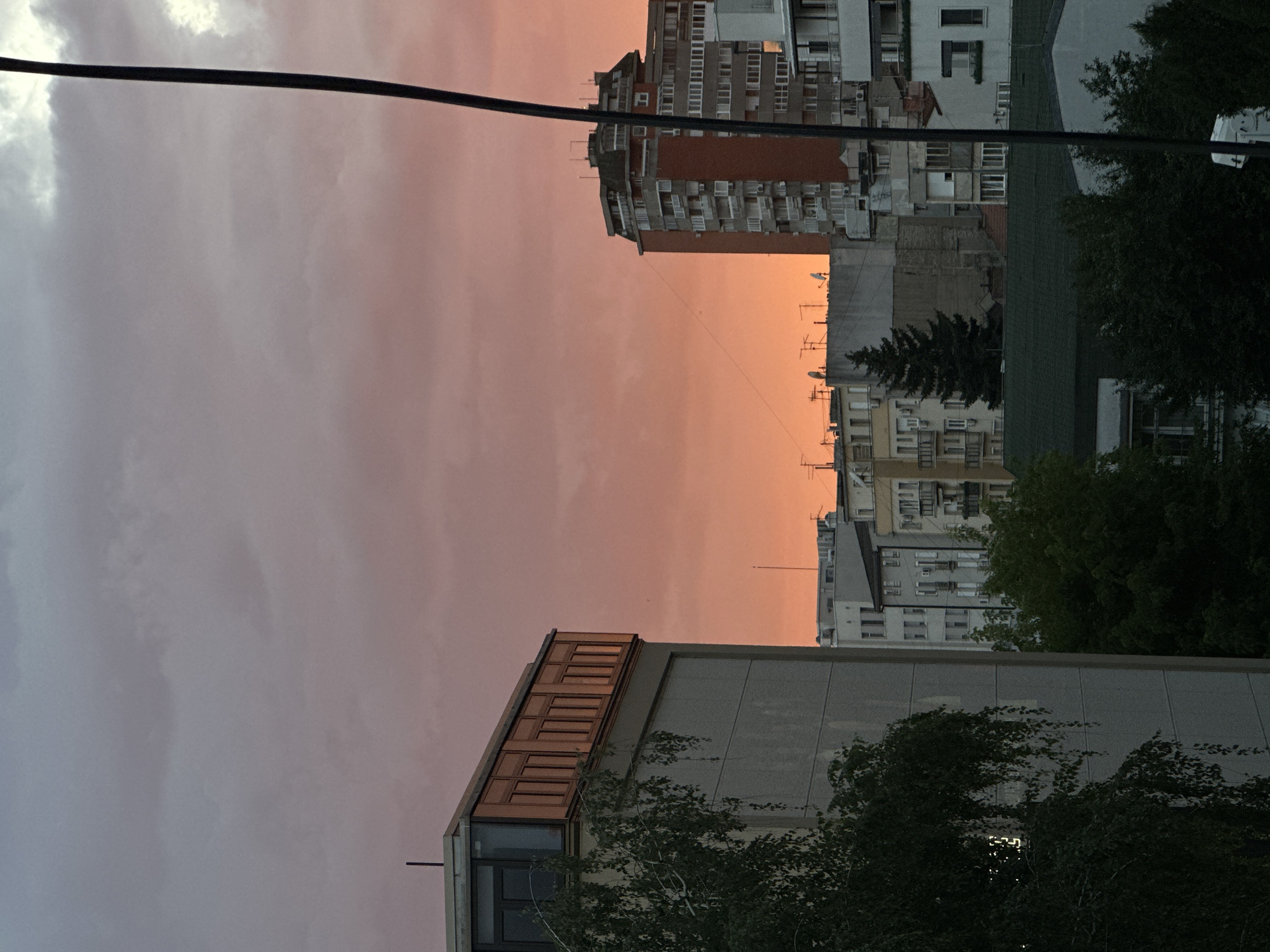 The image captures a twilight cityscape in Vracar Urban Municipality, Serbia, dominated by a warm, orange-pink sky. In the foreground, a portion of a building with a grey facade and a row of wooden-framed windows on its upper level is visible on the left. A weeping willow tree, partially obscuring the building, hangs its branches into the frame from the left side.
Further into the scene, a prominent, tall, reddish-brown apartment building with numerous windows stands out against the sky. To its left and right, other buildings of varying heights and architectural styles are visible, showcasing a typical urban density. Some of these buildings have satellite dishes on their roofs, and balconies are present on several facades.
A dark, vertical line, likely a power line or cable, runs along the right edge of the image, extending from the top to the bottom. In the mid-ground, a tall, dark green evergreen tree is nestled among the buildings. The overall impression is one of a residential urban environment bathed in the soft, fading light of sunset. There are no people or discernible actions depicted in the image.
