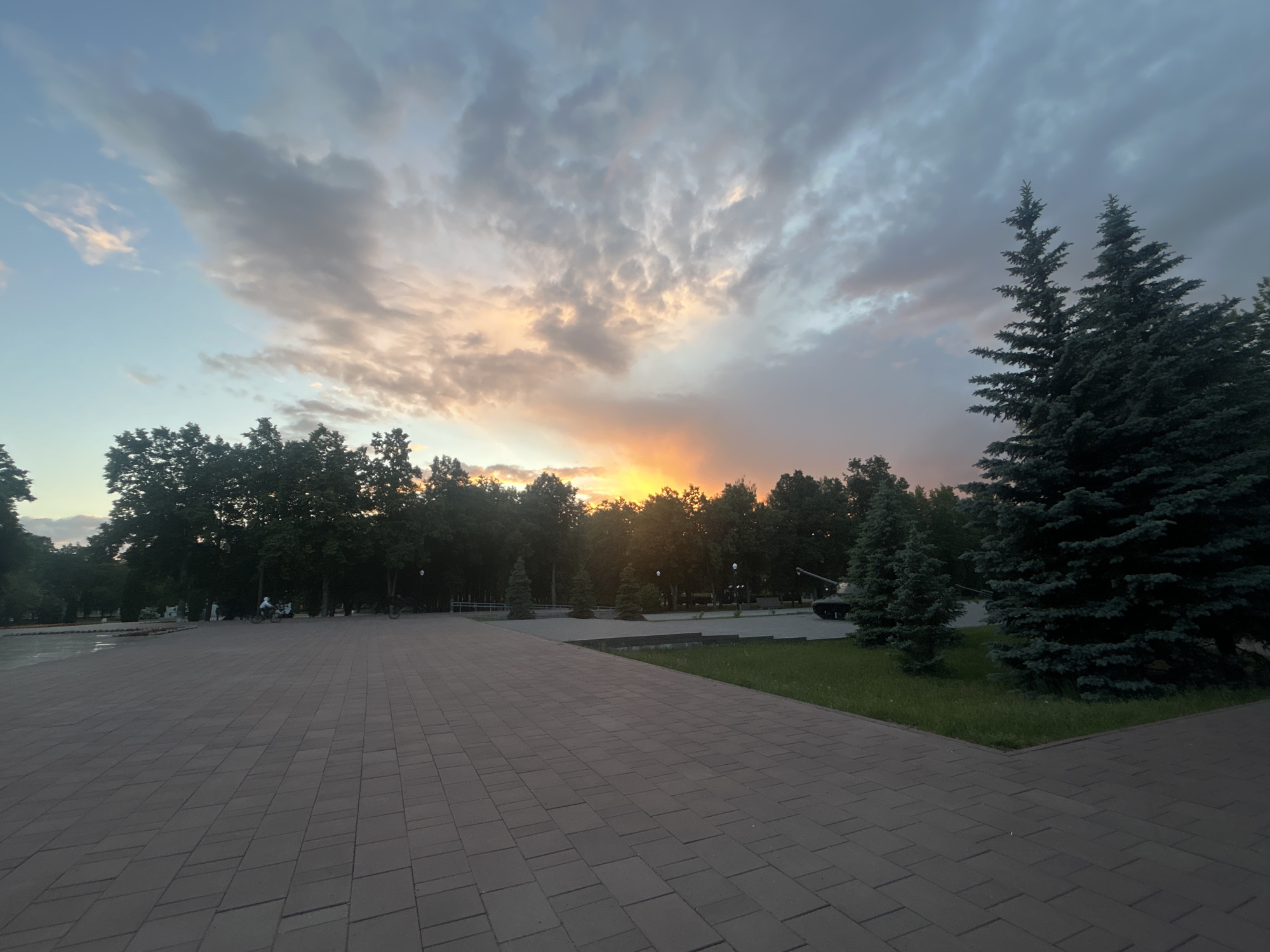 The image depicts a park in Tolyatti, Russia, at sunset. The foreground features a paved area with interlocking bricks. To the right, there are two evergreen trees bordering a grassy patch. In the background, a line of deciduous trees is silhouetted against the setting sun. To the right of the trees, partially obscured by the foliage, is a tank on display. The sky is filled with clouds, tinged with orange and yellow hues from the sunset. In the distance, people are riding bicycles on the left side of the paved road.