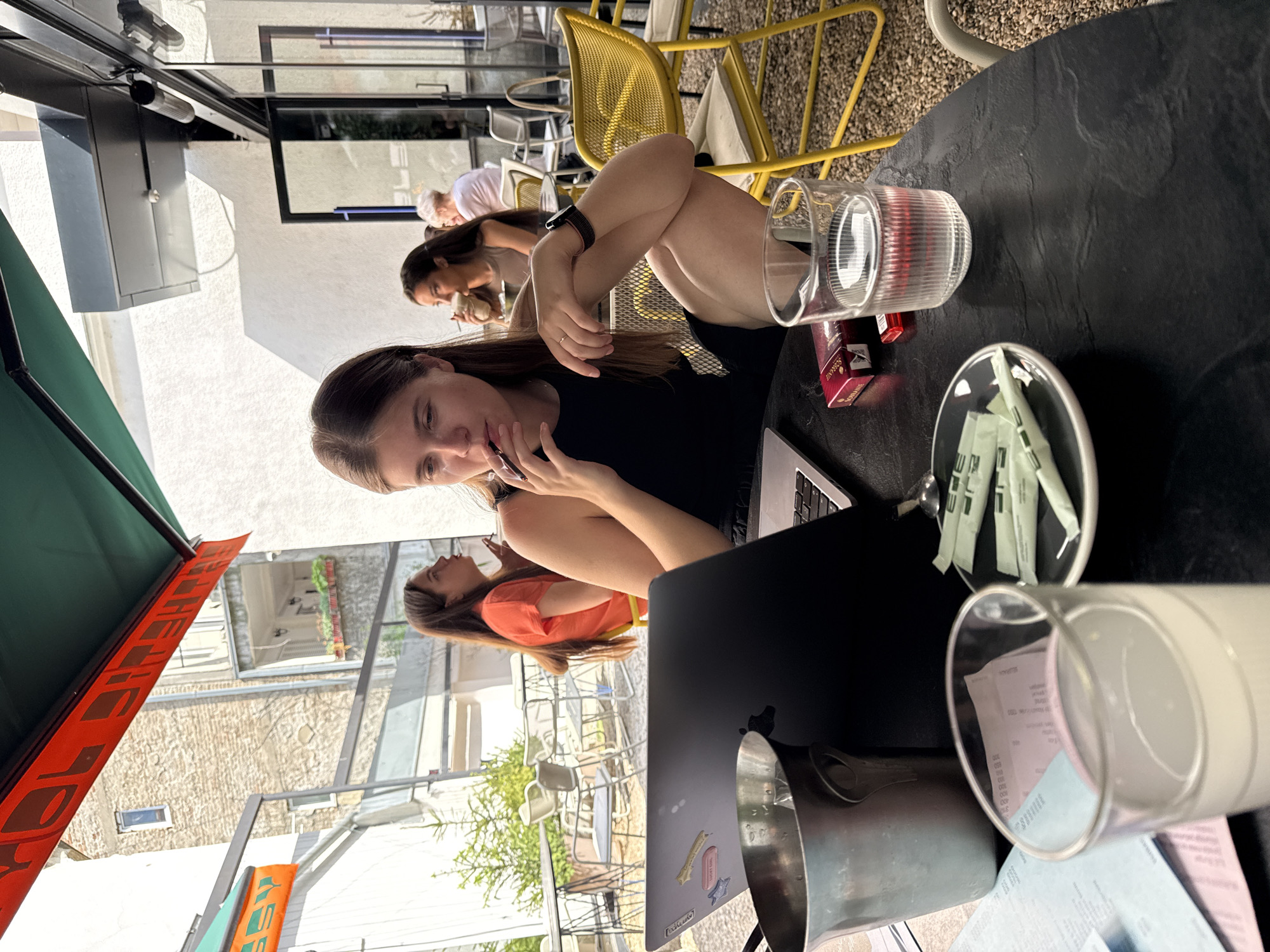 A horizontal, high-angle shot shows a woman seated at a table in an outdoor cafe setting. She is dark-haired and fair-skinned, wearing a black top, and looking at a cell phone she holds near her mouth. Her laptop is open on the table in front of her. At least two other women are seated at nearby tables.
The table holds a glass of water, a pack of cigarettes, a sugar packet holder, receipts in a glass, and a metal pitcher. Outside the cafe, European-style architecture can be seen, possibly in Belgrade, Serbia.