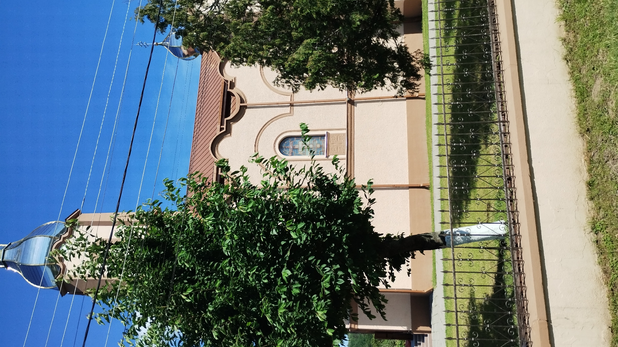 The image depicts an outdoor scene featuring a building with a light brown facade and a brown tiled roof. The building has architectural decorative elements, including curved mouldings above a stained-glass window. A dark metal fence with decorative scrollwork runs along the side of the building, separating it from a grassy area and a concrete walkway. There are trees with lush green foliage in the foreground and to the left of the building. Several power lines are visible against a clear blue sky. The scene appears to be set during the daytime, with bright sunlight casting shadows, suggesting fair weather. The image is oriented vertically. No people or text are visible. The context provided indicates the location is Chynadiiovo, Ukraine.