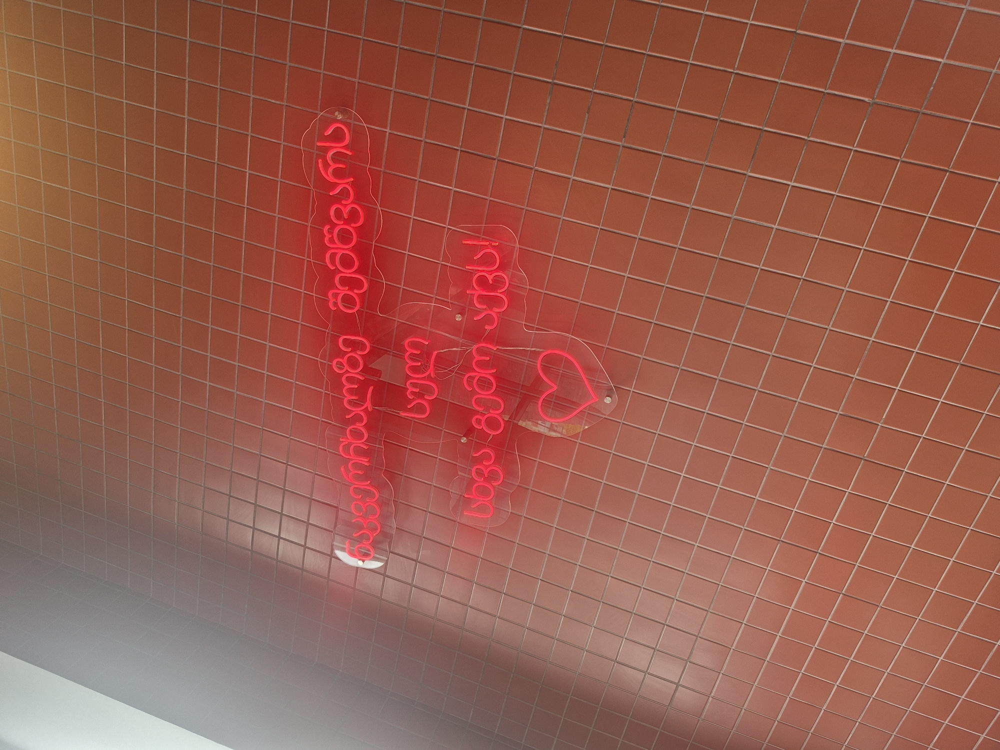 The image is an upward shot showing red neon lettering on a wall covered in small, square orange tiles with light grout. The lettering, which includes a heart shape, is written in Georgian, and the sign is affixed to the wall using clear, curved plastic. The location is likely in Tbilisi, Georgia, given the language used. The perspective suggests the viewer is looking up at the sign.