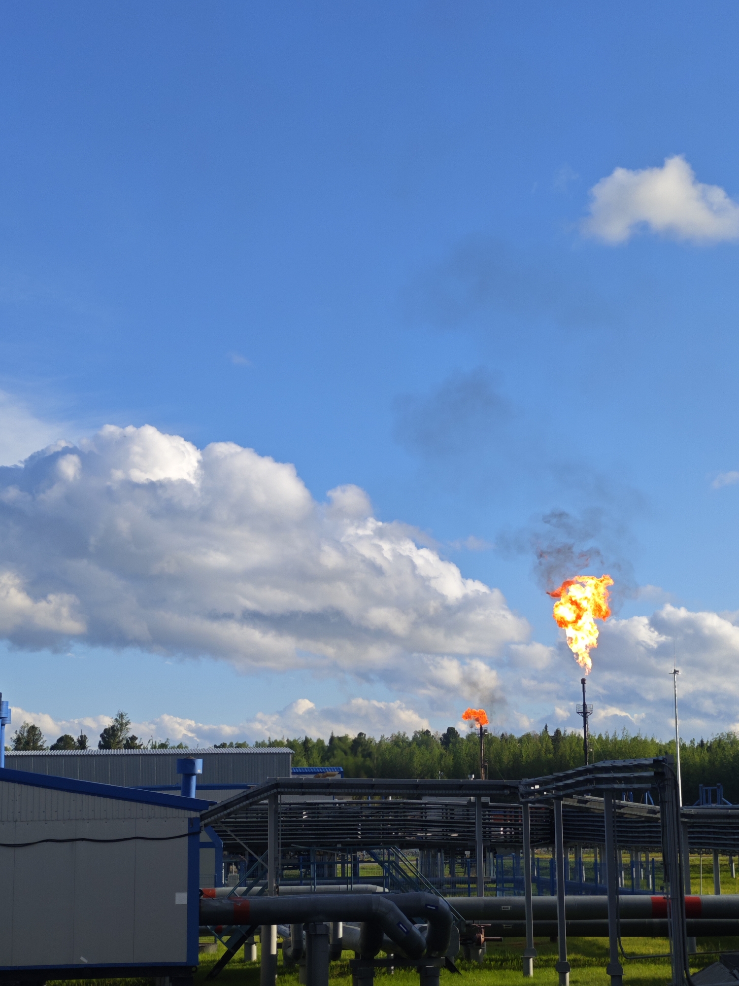 The image shows an industrial facility with prominent flames erupting from two tall flares against a backdrop of blue sky with scattered white clouds. In the foreground, a complex network of pipes and support structures dominates the view. A building with a grey and blue exterior is visible on the left. Beyond the industrial infrastructure, a line of green trees is present. The scene is set in Unknown, Unknown.