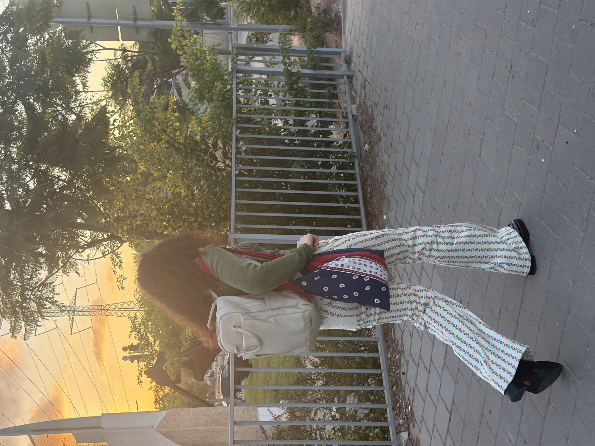 A person walks on a sidewalk with their back to the camera. The person is wearing a green long-sleeved top, a light-colored backpack, and patterned bell-bottom pants with a floral design. They are also carrying a colorful tote bag. In the background, there is a metal fence, trees, and an electrical tower against a sunset sky. The location is the city of Tel-Aviv, Israel, indicated by the presence of the electrical tower, typical of the region, and the overall urban setting.