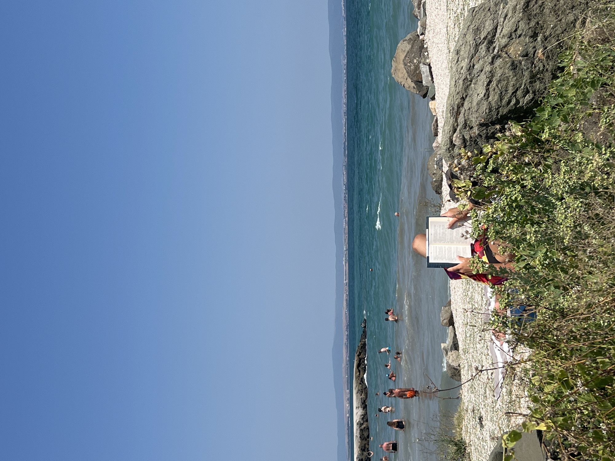 The image depicts a scene at the beach in Pomorie, Bulgaria, under a clear blue sky. The foreground is dominated by lush green vegetation and large rocks. A person is seated in the mid-ground, partially obscured by foliage, engrossed in reading a book. This individual is wearing a colorful sarong and a bikini.
Beyond the beach, the turquoise waters of the sea are populated by numerous people swimming and enjoying the water. The sea extends to the horizon, where a faint outline of land with distant buildings and hills is visible. A stone breakwater extends into the sea from the left side of the image, with more people visible near and on it. The beach itself appears to be covered in small white pebbles. The overall atmosphere is one of a sunny, leisurely day at the seaside.