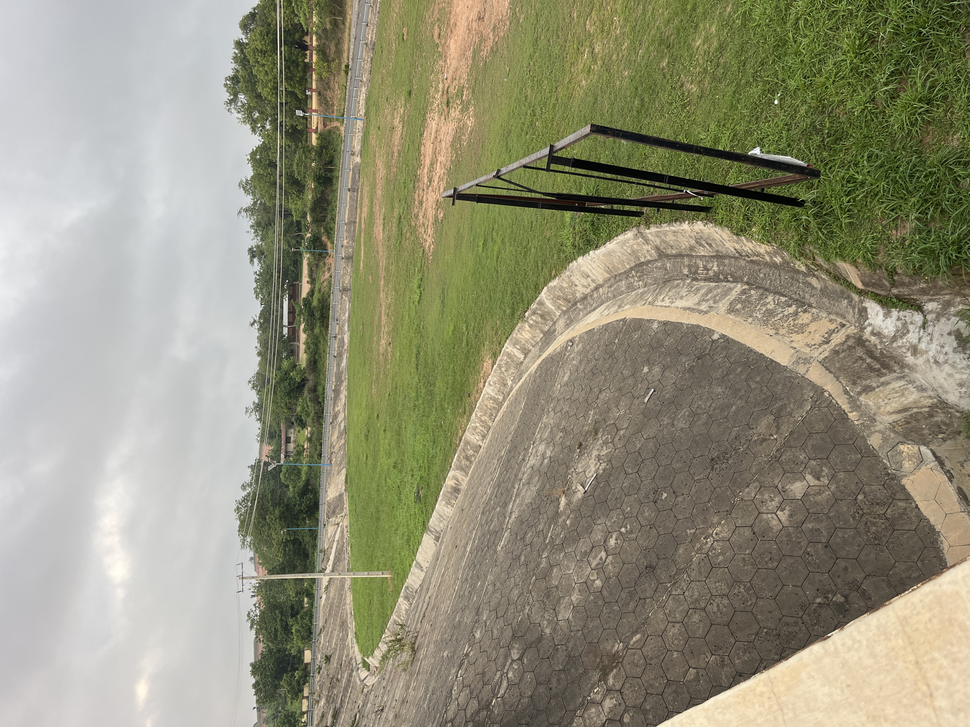 Here is a description of the image:
The image shows a view of a concrete walkway or track with a hexagonal pattern, alongside a grassy field. A black metal structure stands partially on the grass. In the background, trees line the horizon under a cloudy sky. Based on the mention of Osogbo, Nigeria, this is likely a recreational or sports ground in that city. No people are visible in the image.
