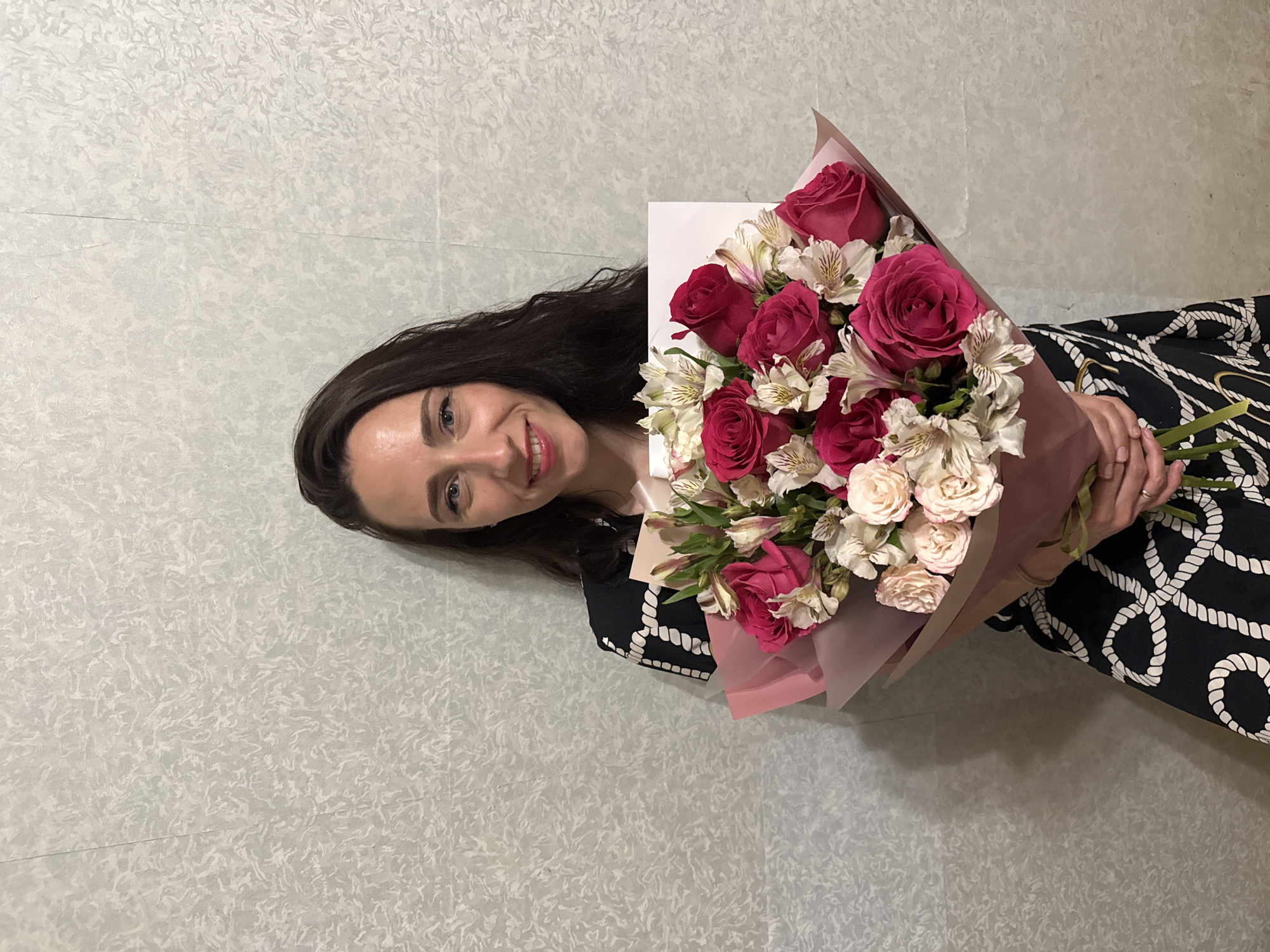 A woman with long dark hair, dressed in a black and white patterned dress, is holding a bouquet of flowers. The bouquet contains red and white roses, along with white alstroemeria. The background appears to be a textured wall with a light gray and white pattern. The location of the scene is not identified as Saint Petersburg, Russia.