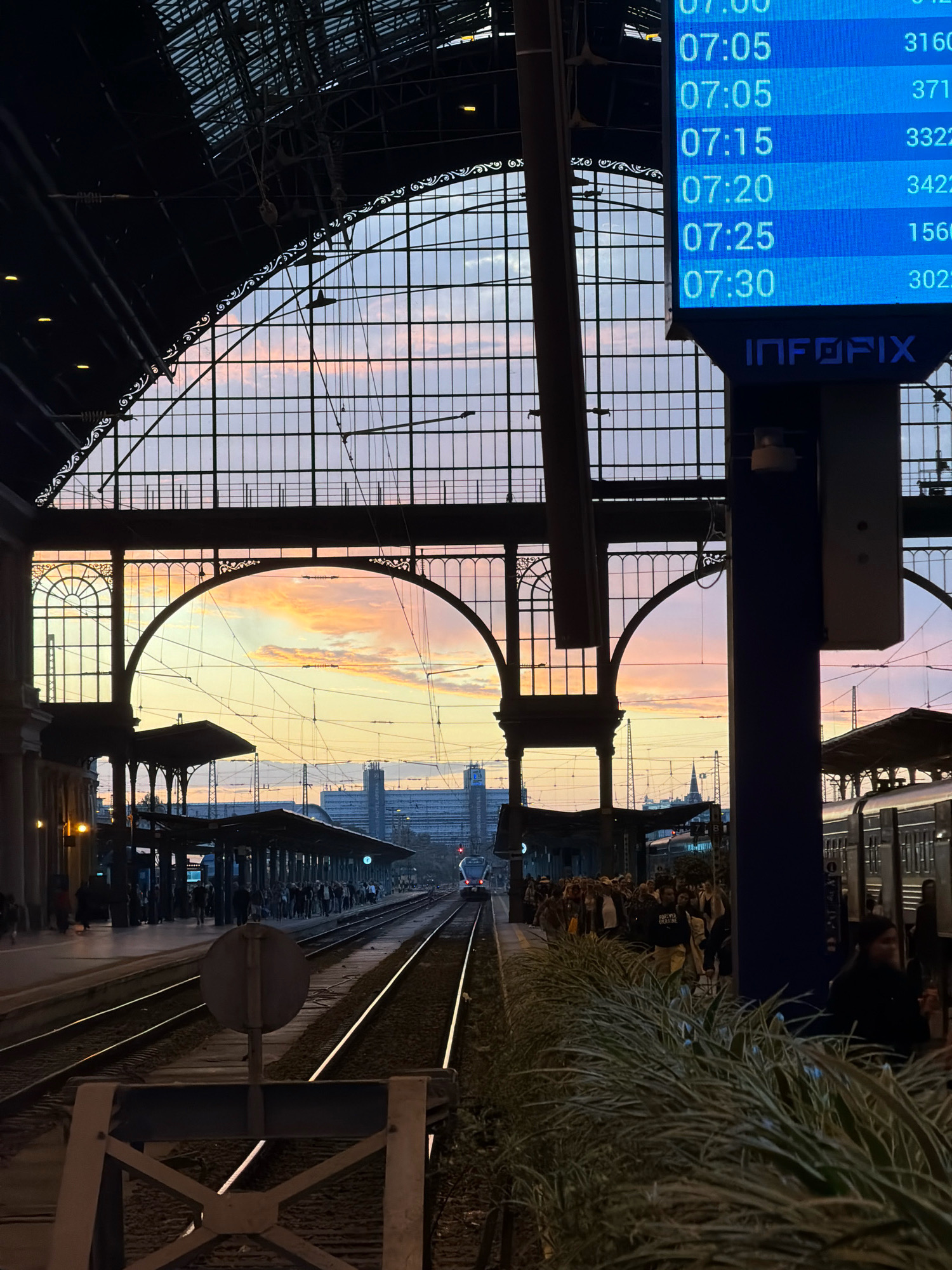 This is a low-angle, medium shot of a train station in Budapest, Hungary at dusk. The image is dominated by a large, arched glass ceiling with intricate metalwork. Through the glass, a colorful sunset sky with pink and orange clouds is visible. In the foreground, train tracks run into the distance, with a train approaching on the right.
On the left side of the image, a platform is visible with people waiting. A large digital display board shows train departure times and destinations, with the times ranging from 07:00 to 07:30 and destinations such as 3160, 371, 3322, 3422, 1560, and 3022. The brand "INFOFIX" is displayed on a dark blue pillar.
The architecture of the station is grand and industrial, with arched doorways and a complex network of overhead wires for the trains. The overall atmosphere is one of transition, with the fading light of day and the bustle of evening travel.
