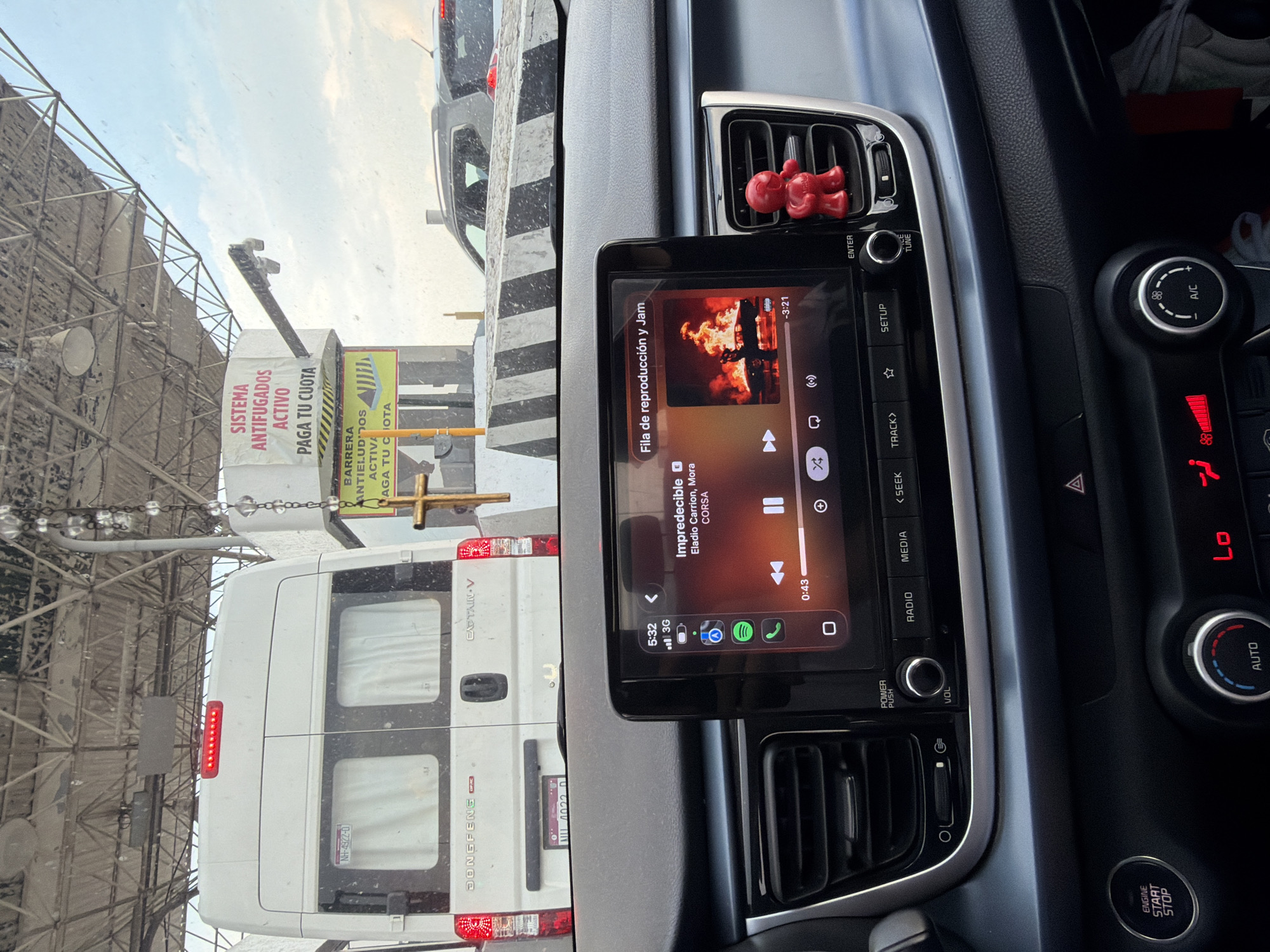 The image shows the interior of a car, specifically the dashboard area. The infotainment system is displaying a music playlist, featuring the song "Impredecible" by Eladio Carrion and Mora. The car's climate control is set to 88 degrees. There's a red car air freshener above the screen, and the engine start/stop button is visible.
Through the windshield, we see a toll booth with signage in Spanish, indicating "SISTEMA ANTIFUGADOS ACTIVO" and "PAGA TU CUOTA". A white van, marked "DONGFENG CAPTAIN V" with license plate "NH-4922-0" is visible in front of the car.
Based on the road signs and the vehicle registration, the location is likely a toll road in Mexico. Based on other data points, the city is identified as Metepec, Mexico.