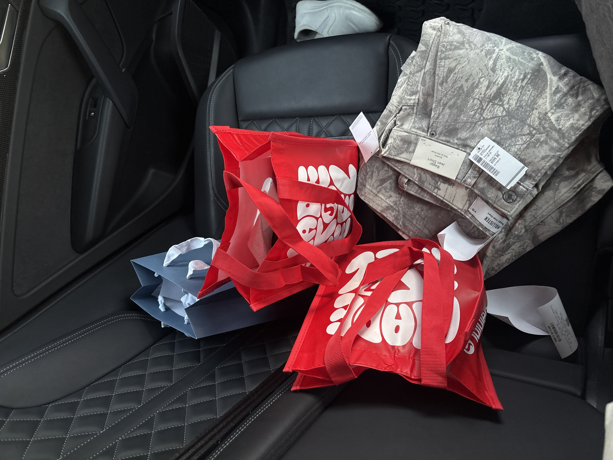 The image displays the interior of a car, specifically the back passenger seat, laden with recently purchased items. The car features black leather seats with prominent white diamond-patterned stitching on the seat bases and a dark door panel on the left.
In the foreground, two vibrant red reusable shopping bags with white stylized logos, possibly reading "JUJUB" or "JUJUBA", are prominently placed. Behind them, a blue paper bag and a partially visible white paper bag are also present. On the right side of the seat, a folded pair of "HOLLISTER Baggy Jean Short" is visible, sporting a gray and white abstract or camouflage pattern. Attached tags indicate size "w30," "Stretch," and "Hits at the Knee." One tag also shows a price of "$599.00" along with "PRECIO ORIGINAL" and a barcode, suggesting Mexican Pesos given the context of San Bernardino Tlaxcalancingo, Mexico. Further back on the rear seat, a light-colored (possibly white or light gray) shoe is resting near the headrest. Several receipts or paper tags are scattered among the items. The scene suggests a recent shopping trip, with the purchases being transported in the car.