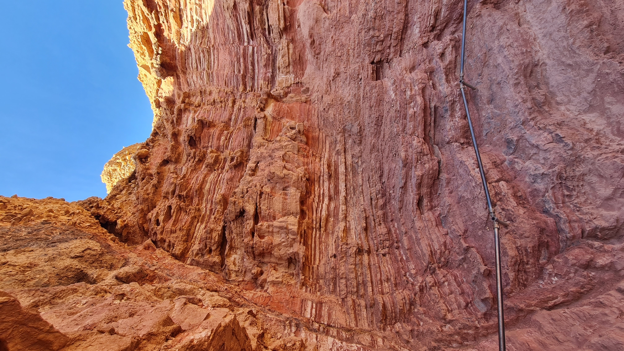This photograph shows a close-up of a rust-colored rock formation with a mostly clear blue sky visible in the top left corner. The rock formation displays vertical striations, and there is a metal pipe with connectors attached to the rock face. The location is identified as being in the city of Hevel Eilot Regional Council, Israel. The scene does not contain any people, but the presence of the pipe suggests human interaction or alteration to the natural landscape.