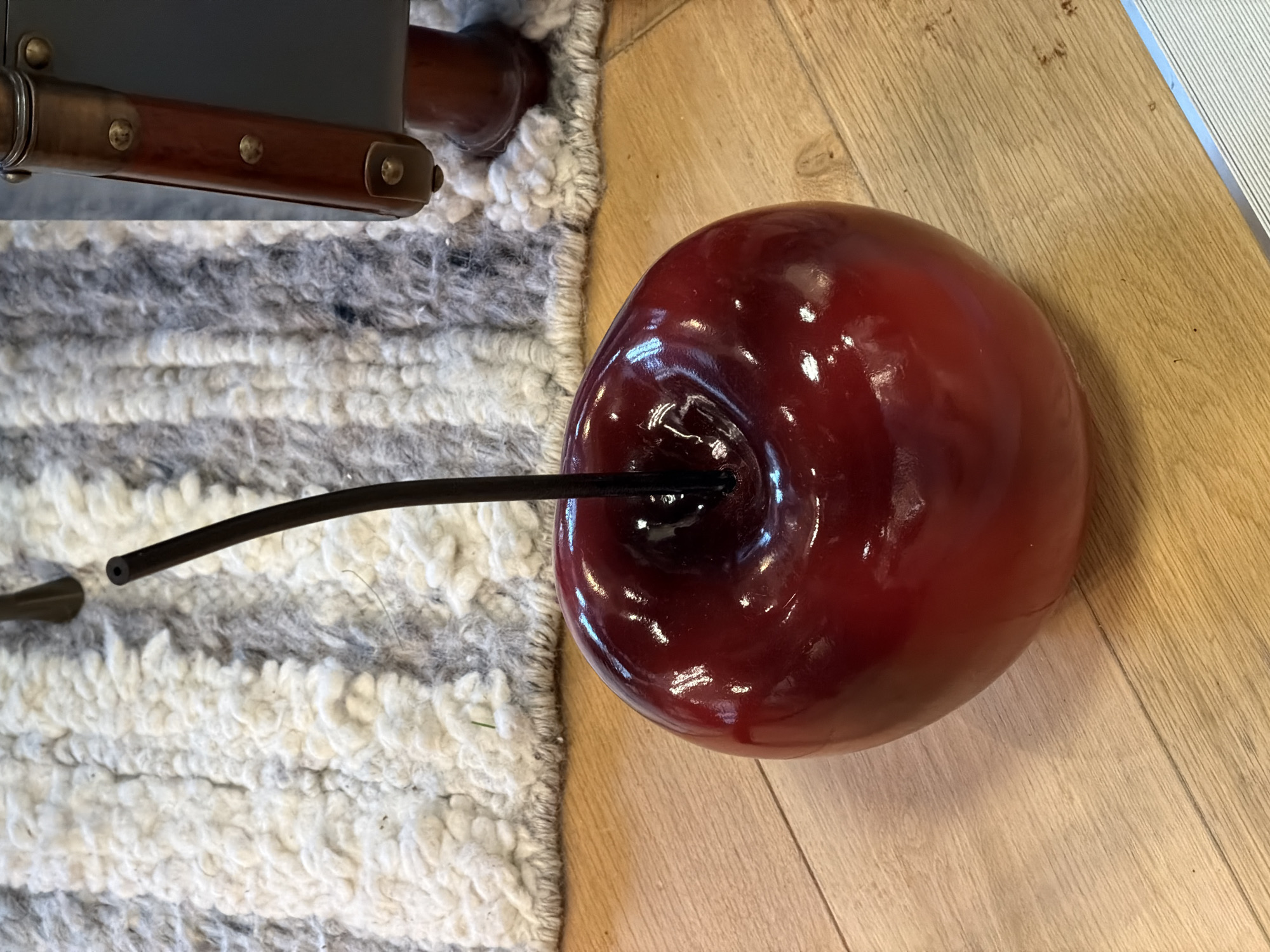The image depicts a shiny, red, cherry-shaped object with a black stem protruding from it, resting partially on a beige wooden floor and partially on a gray and white striped rug. An indistinct piece of furniture is visible in the upper left corner of the frame. There is no indication of the location.
