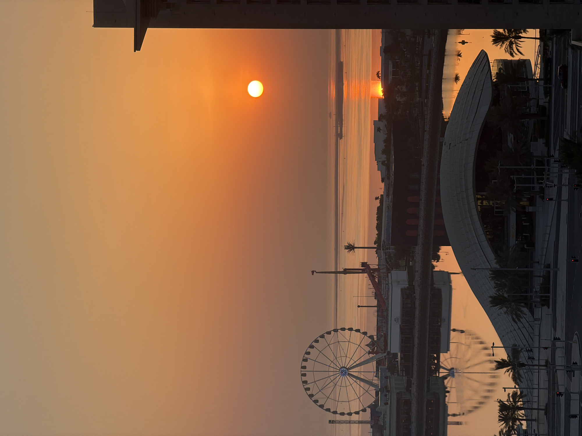 This is a vertical, eye-level shot of a sunset over the waterfront in Lusail, Qatar. The sky is filled with a gradient of orange and yellow hues, with the sun a bright, glowing orb low on the horizon, casting a shimmering reflection on the calm water. In the foreground, the distinctive curved roof of a modern building, possibly a stadium or exhibition center, is visible, with palm trees lining its perimeter. Further in the distance, a Ferris wheel stands tall, illuminated with lights, suggesting an amusement park or entertainment district. Various other structures, including buildings and possibly a roller coaster, are scattered along the waterfront. A large cargo ship is visible in the middle distance, its reflection also visible on the water. The scene is characterized by the warm, soft light of sunset, creating a serene and picturesque urban waterfront landscape. There are no people visible in the image.