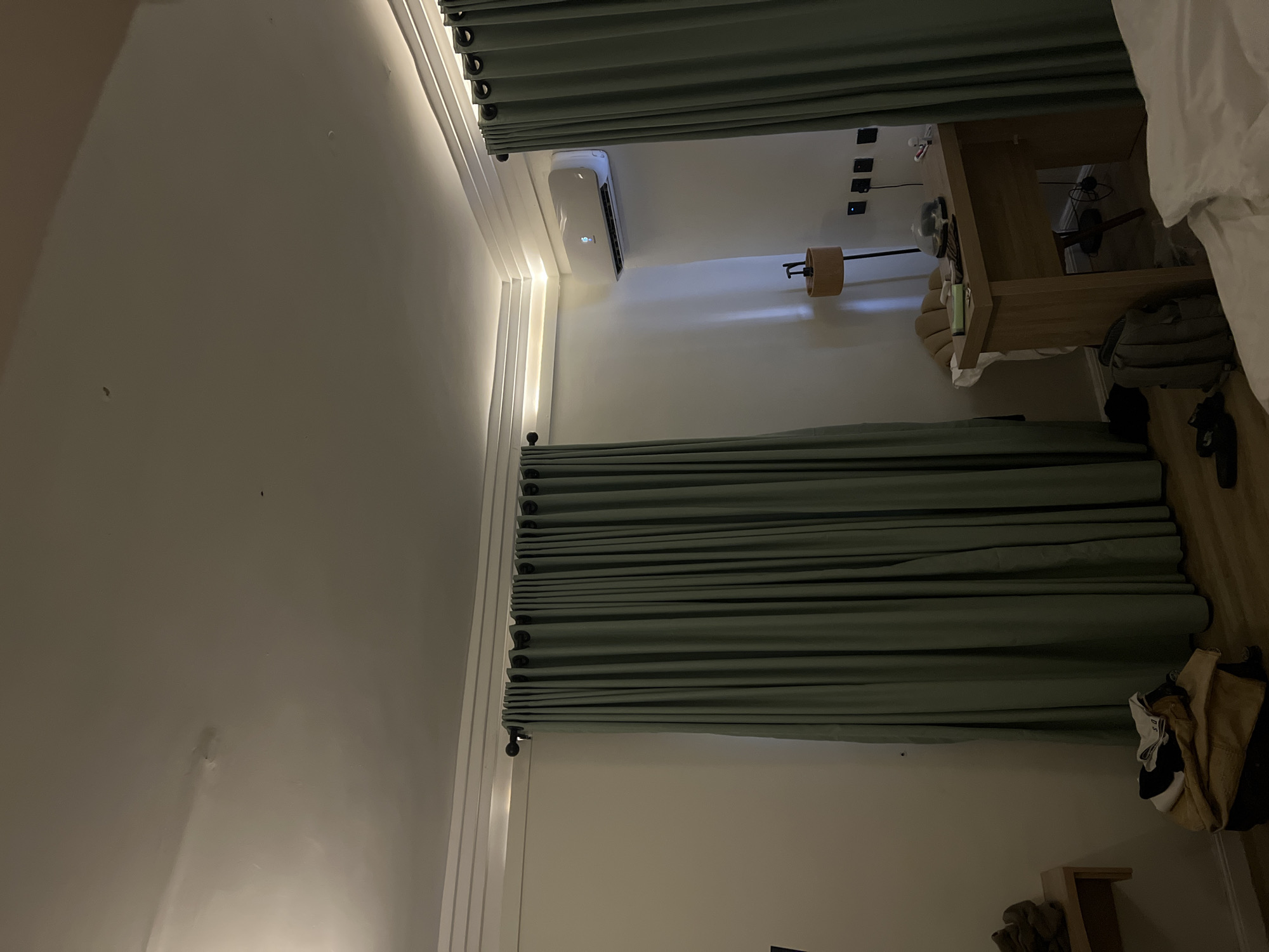 The image displays an indoor room, likely a bedroom or hotel room, photographed in a portrait orientation but rotated 90 degrees counter-clockwise, making the walls appear as ceiling and floor. The room features a modern design with light-colored, possibly off-white, walls.
Two sets of pleated, light green (or teal) curtains are drawn, covering what would be windows if the image were correctly oriented. Recessed strip lighting runs along the junction of a wall and the ceiling (appearing as the left wall in the rotated image), casting a warm, ambient glow.
A white split-unit air conditioner is mounted high on one wall, displaying a digital reading of "25°C". Below it, to the right, is a wall-mounted lamp with a cylindrical wooden shade and a black arm. Further right, a wooden bedside table or small desk is visible, with a few indistinct items placed on it. Above this furniture, several black wall outlets or switches are mounted on the wall. A white bed with a duvet is partially visible in the upper right corner of the frame. A green or khaki-colored backpack is seen near the bottom right, next to some folded clothes or fabric items.
The room is dimly lit by artificial light, suggesting it is evening or night, or that natural light is completely blocked by the curtains. The overall impression is one of a functional and contemporary space. No specific activity is taking place, and no people are visible. The scene is located in Gwarinpa, Nigeria.