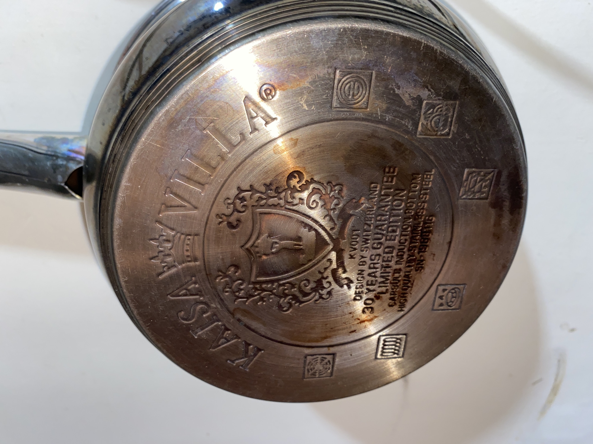 The image shows the bottom of a stainless steel saucepan. Circular text around the edge reads "KALSA VILLA". In the center is a heraldic shield design. Text below the shield states "KV001 DESIGN BY SWITZERLAND 30 YEARS GUARANTEE LIMITED EDITION CAPSULTE INDUCTION BOTTOM HIGH QUALITY STAINLESS STEEL SN-1980118". The pot appears well-used with discoloration and surface scratches.