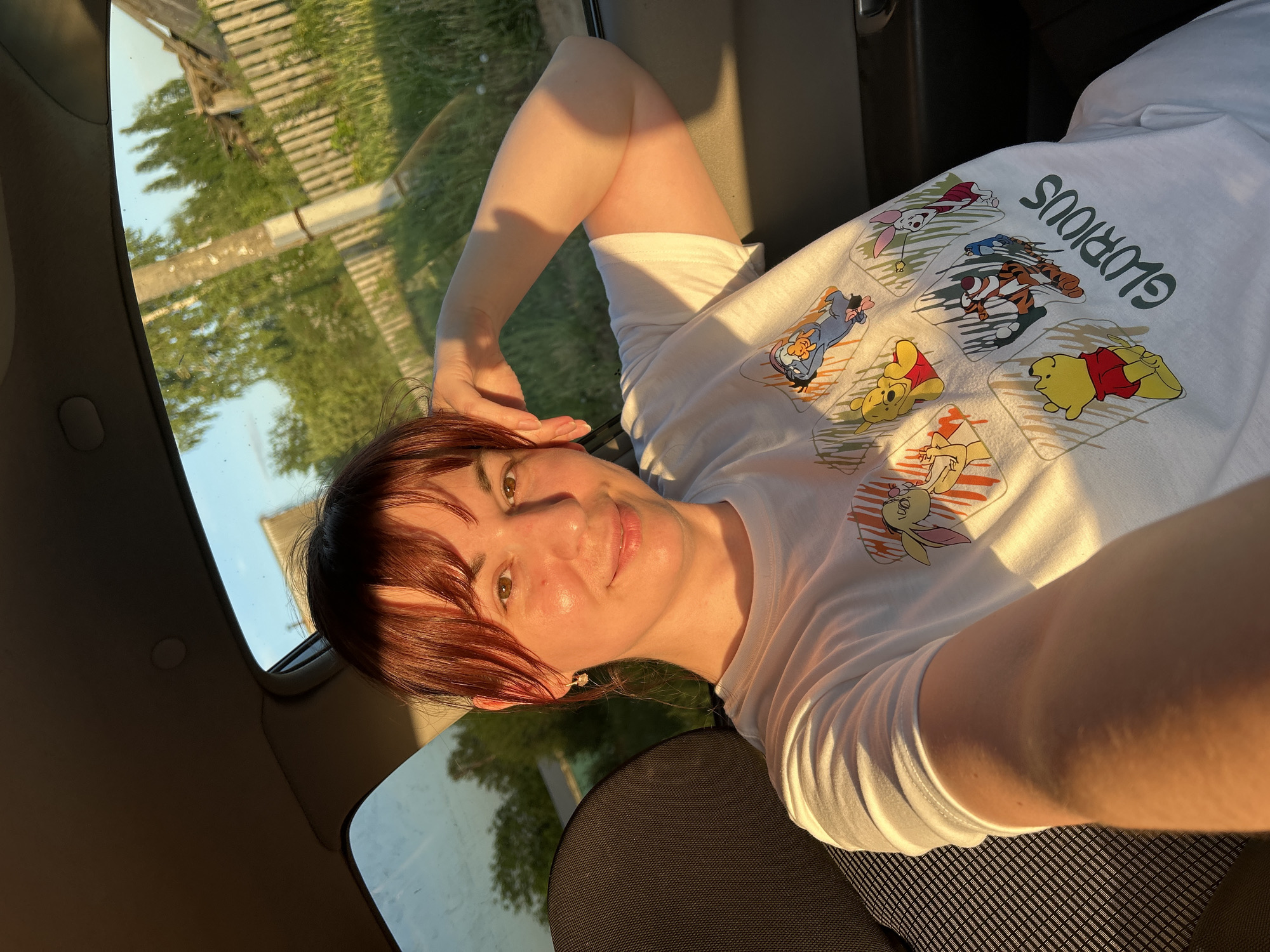 A fair-skinned woman with short auburn hair is lying down in what appears to be a car, taking a selfie. She is wearing a white t-shirt printed with Winnie the Pooh characters and the word "GLORIOUS". The sun is shining, casting shadows across her face and clothing. Outside the car window, trees and part of a wooden fence can be seen. The seat has a black and gray striped texture. While there is no direct evidence of being in Фоки, Russia from the image itself, the prompt states to assume the location is Фоки, Russia.