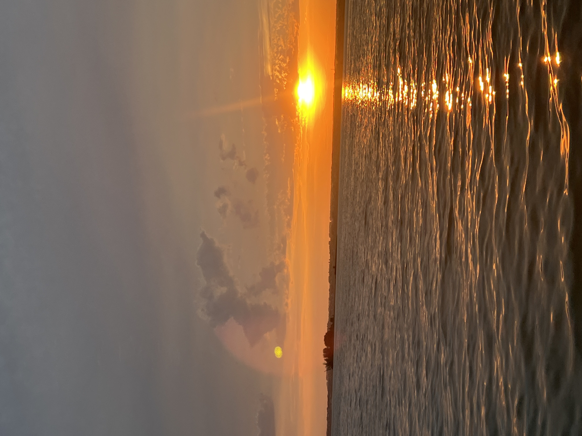This image depicts a sunset over a body of water, likely the Black Sea, near Beregovoe, Russia. The sky is a gradient of colors, with deep grays and blues at the top transitioning to vibrant oranges and yellows near the horizon. The sun is a bright, concentrated orb, casting a shimmering reflection on the rippling water. In the distance, a faint silhouette of land is visible, dotted with some trees. There are no people or specific objects that indicate any activity. The overall scene is serene and natural.
