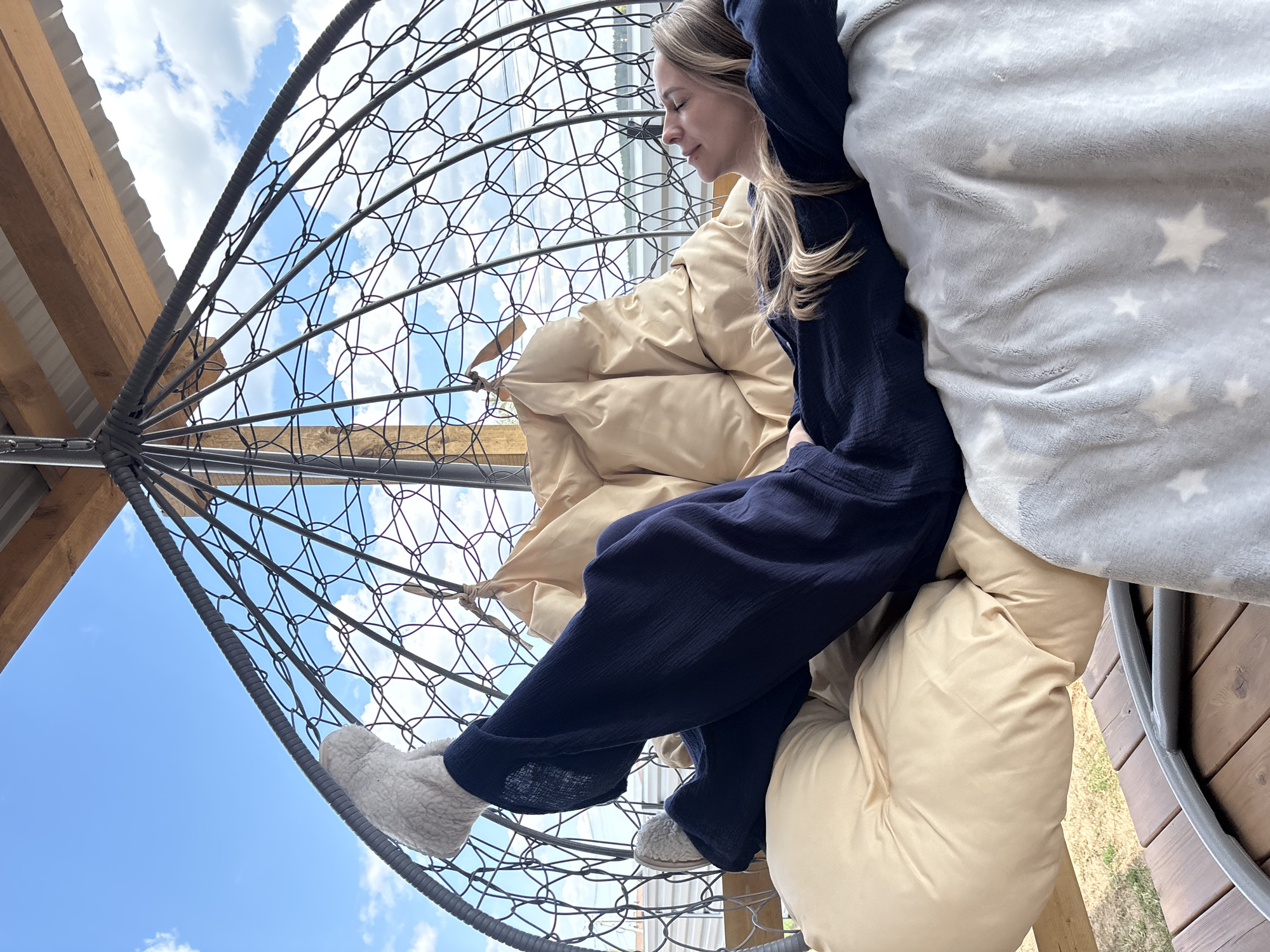 A woman sits in a spherical hanging chair on a wooden deck, partially obscured by cushions and a blanket. She wears a dark blue outfit and light-colored slippers. The chair is constructed of a dark-colored metal framework against a backdrop of blue sky with scattered clouds. A portion of a building's roof structure is visible to the left. The location is Chaikovsky, Russia.