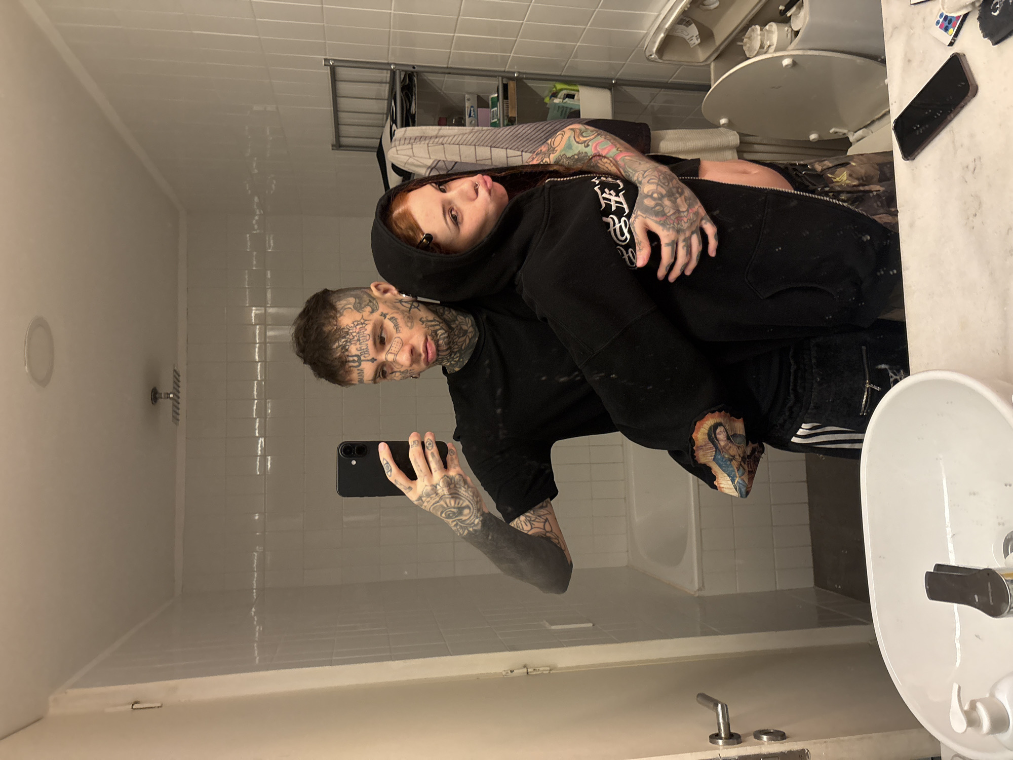 The image shows two individuals posing for a selfie in a bathroom. The person taking the photo appears to be male, heavily tattooed on his face and arms. He is holding a black smartphone. A woman with red hair is leaning against him, also tattooed. The bathroom has white tiled walls, a white bathtub, a toilet, and a sink. There are items on the countertop including what looks like phone, makeup and remote control. The location is not identifiable as Buenos Aires, Argentina, based solely on the image.