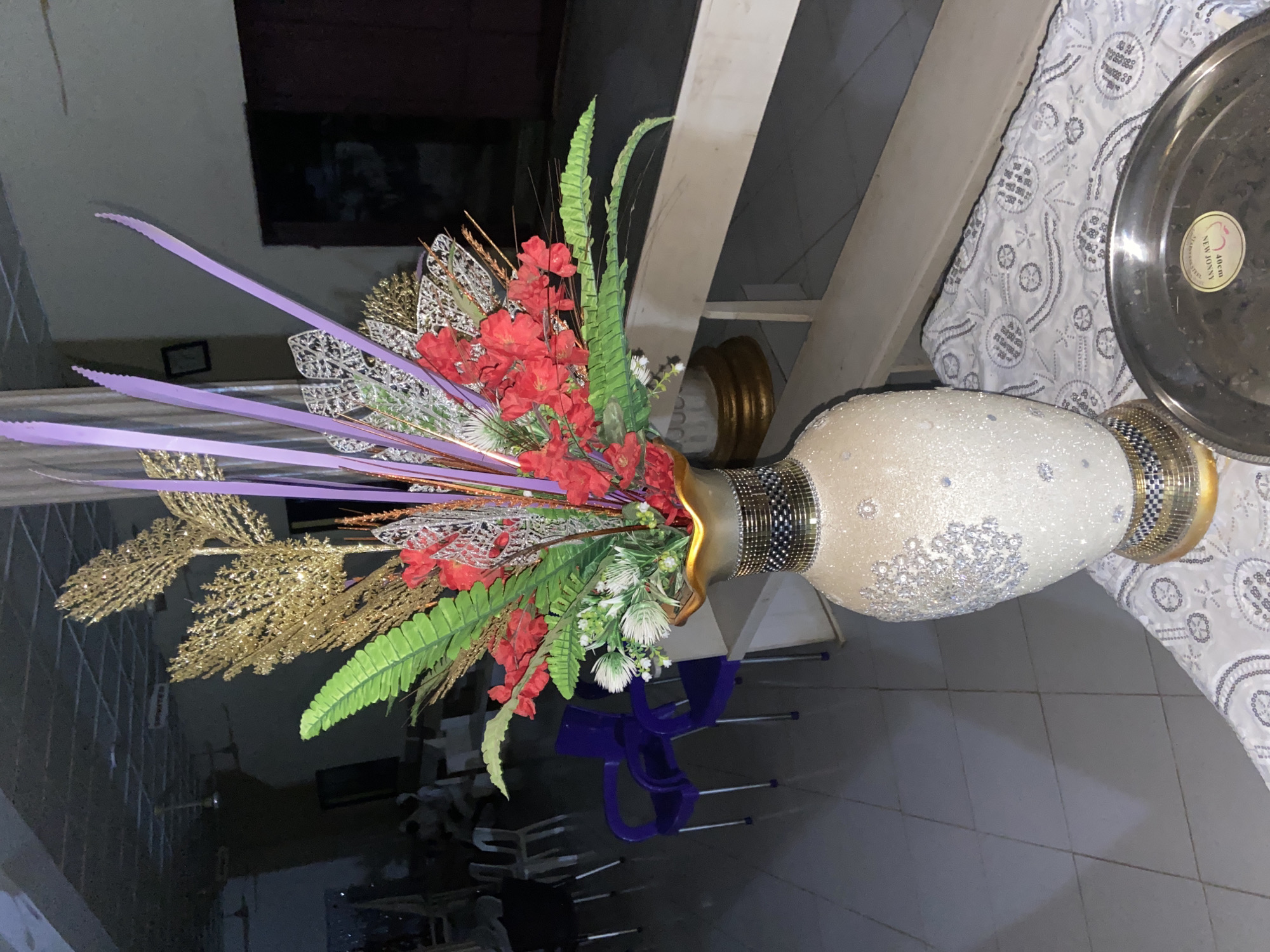 The image shows a decorative floral arrangement in a large, ornate vase. The vase is textured and adorned with small sparkly elements, and has a metallic rim. The floral arrangement itself is elaborate, featuring red artificial flowers, green artificial leaves, and a mix of gold and silver decorative foliage. There are also long, flat, purple ribbons extending from the arrangement. The arrangement is placed on what appears to be a table covered with a patterned white tablecloth, on which rests a metallic platter. The background is dimly lit, showing a wall and some furniture, including chairs that appear to be stacked. The scene is indoors, likely in a domestic setting. The image is taken at an angle, with the vase and arrangement dominating the frame. No people are visible in the image.
There is some visible text on a sticker on the metallic platter. The text appears to be in a circular format and includes "NEW JENNY" and "40 cm". The overall impression is one of a formal or decorative display, possibly for an event or as a permanent fixture in a home. The lighting suggests it could be evening or a room with limited natural light. Given the location context of Lagos, Nigeria, the style of the decoration might reflect local aesthetics.