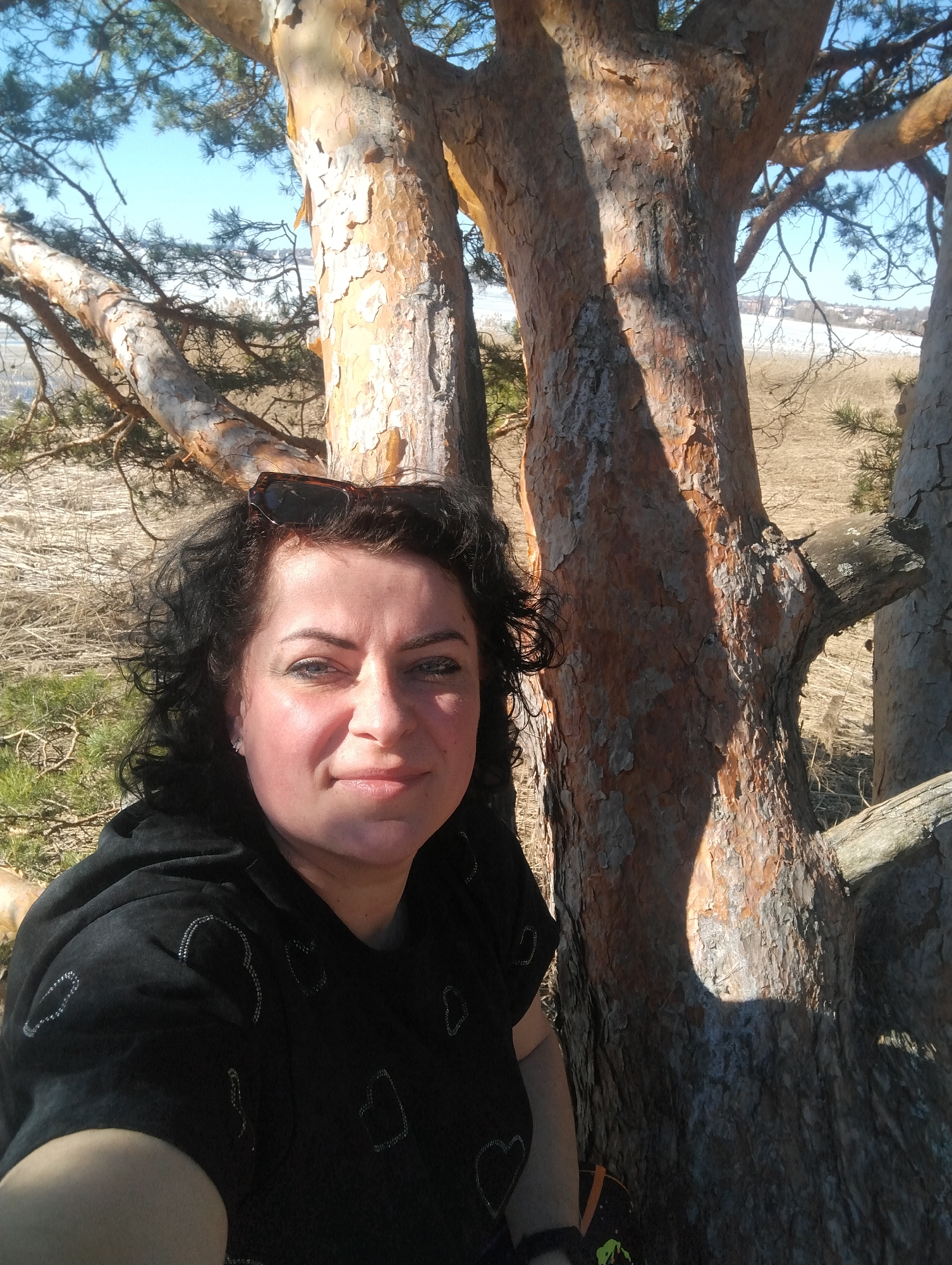 A selfie taken in daylight in a park in Votkinsk, Russia. The subject, a middle-aged woman with dark, curly hair and sunglasses perched atop her head, is wearing a dark, heart-patterned top. She is positioned close to a tree with peeling bark, its branches extending toward the sky. In the background, dry vegetation and a partially frozen body of water are visible, with a cityscape on the horizon.