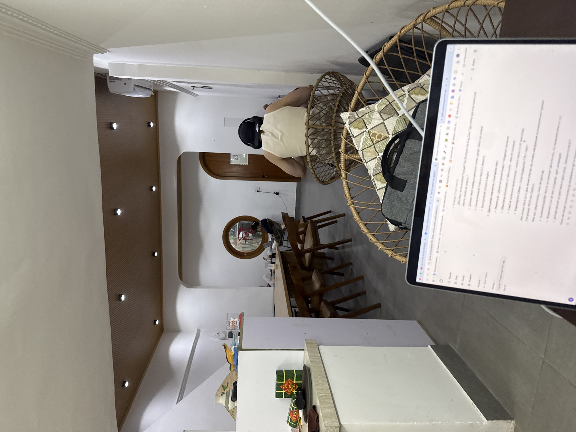 Here is a concise description of the image:
The image shows an interior shot of a room in Vietnam, likely in Đà Nẵng. The view is from a low angle, looking slightly upward.
A person wearing a VR headset and a light-colored tank top is seated in a woven chair, with a laptop in the foreground partially obscuring the view. The laptop screen displays text, presumably work-related. In the background, there is a dining area with a long table and chairs, and a round mirror hanging on the wall. A kitchen area is visible, with white cabinets and some food items. The lighting is warm, and the overall environment appears to be a modern, comfortable living space.