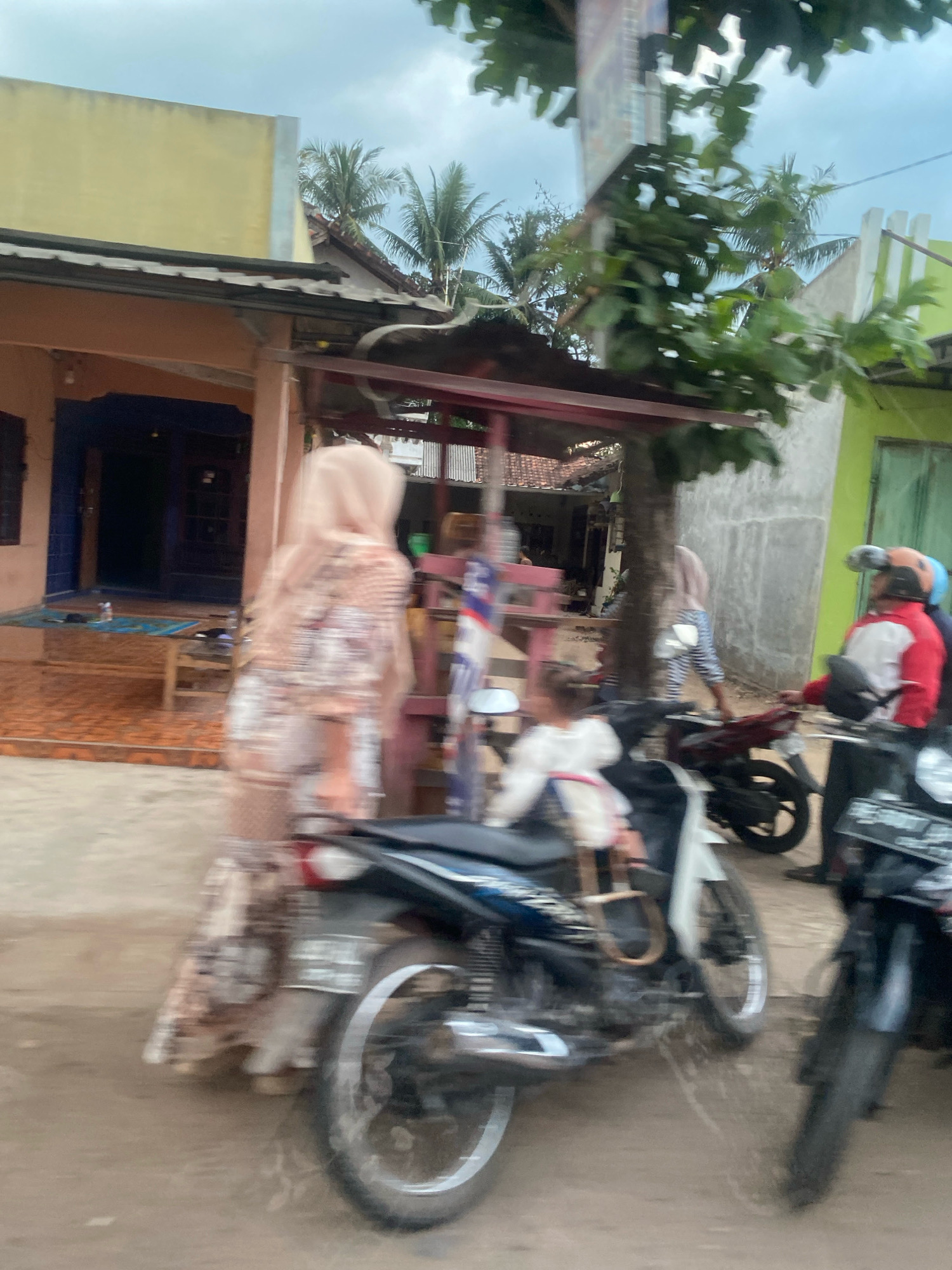 A slightly blurry, outdoor scene in Toto Katon, Indonesia shows a dirt or unpaved road in front of two buildings. The building on the left is light yellow and has a dark blue door. A wooden bench is in front of it. The building on the right is light green. Several people and motorcycles are visible. A person wearing a light-colored headscarf and patterned clothing stands near the yellow building, facing away from the camera. A child in a white shirt is seated on a motorcycle. Another motorcycle is parked to the right, and a person wearing a red and white jacket and helmet is on it. In the background, palm trees are visible. The sky is overcast.
