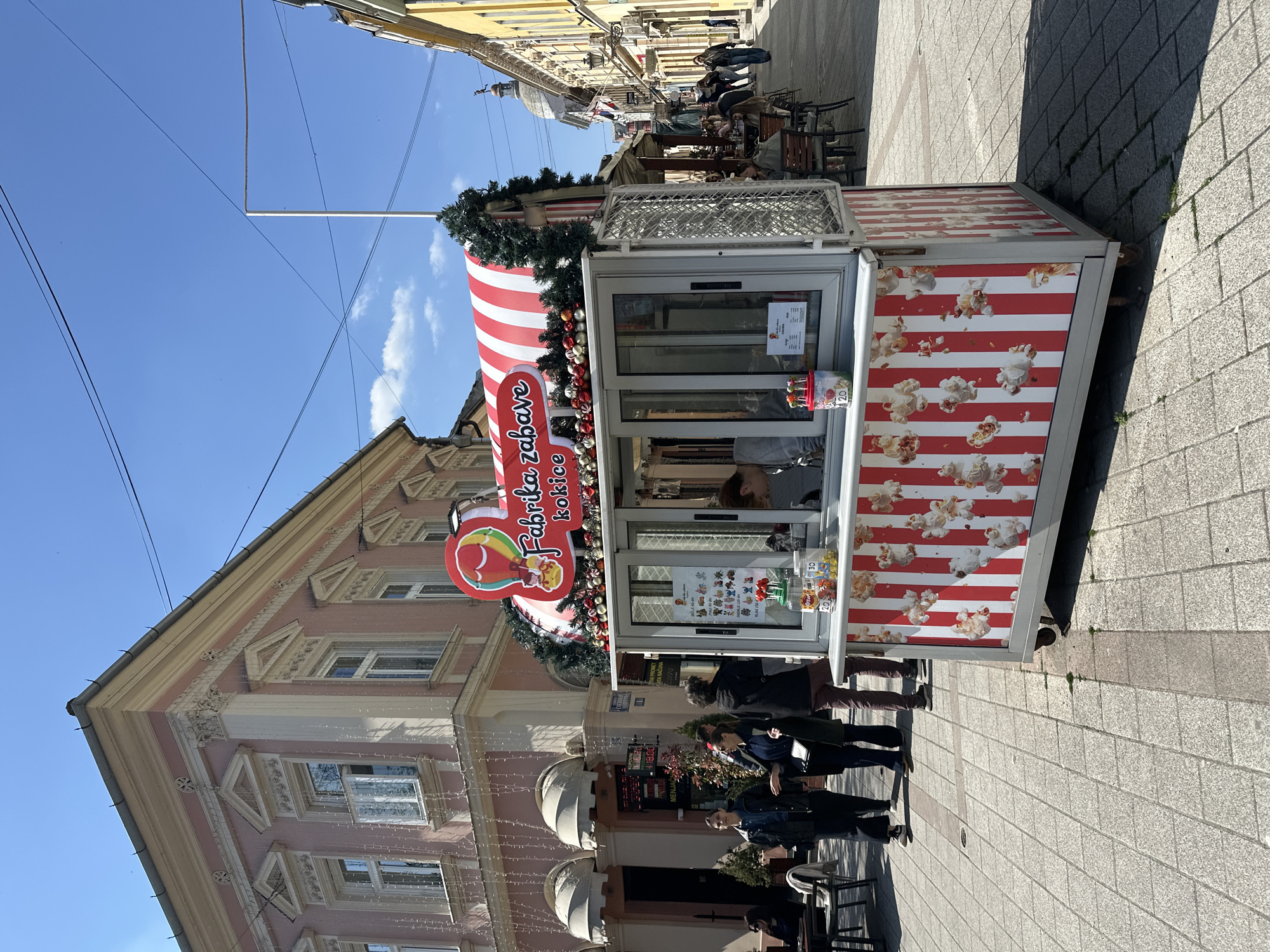 The image shows a view of a street scene in Novi Sad, Serbia, featuring a decorated "Fabrika kokica" (popcorn factory) kiosk. The kiosk is adorned with red and white stripes, Christmas decorations, and popcorn graphics.
Several people are standing and walking around the kiosk area. The surrounding environment includes buildings, some decorated with exterior lighting, and outdoor seating for cafes or restaurants. The street is paved with light-colored stones.
