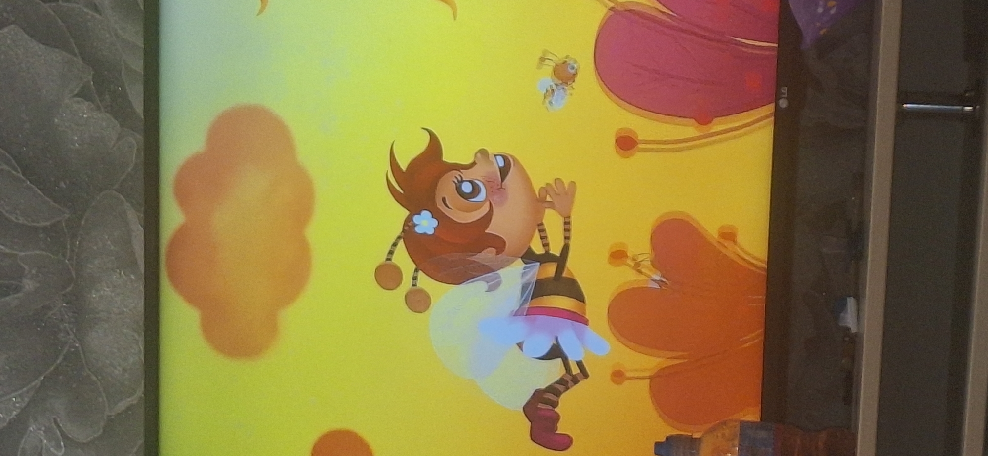 This is a close-up, vertical, eye-level shot of a television screen displaying a cartoon. The screen shows a bee character with brown hair, large eyes, and a striped body, wearing a yellow and white translucent wing structure. The bee is in mid-flight against a bright yellow background with an orange cloud formation at the top. There is a smaller bee character in flight to the right of the main character. In the foreground, at the bottom of the screen, are stylized floral elements in orange and pink, with a bottle of liquid on the left. The television is on a dark grey shelf, with a textured, dark grey surface above it. There is no identifiable location in Bila Tserkva, Ukraine visible.