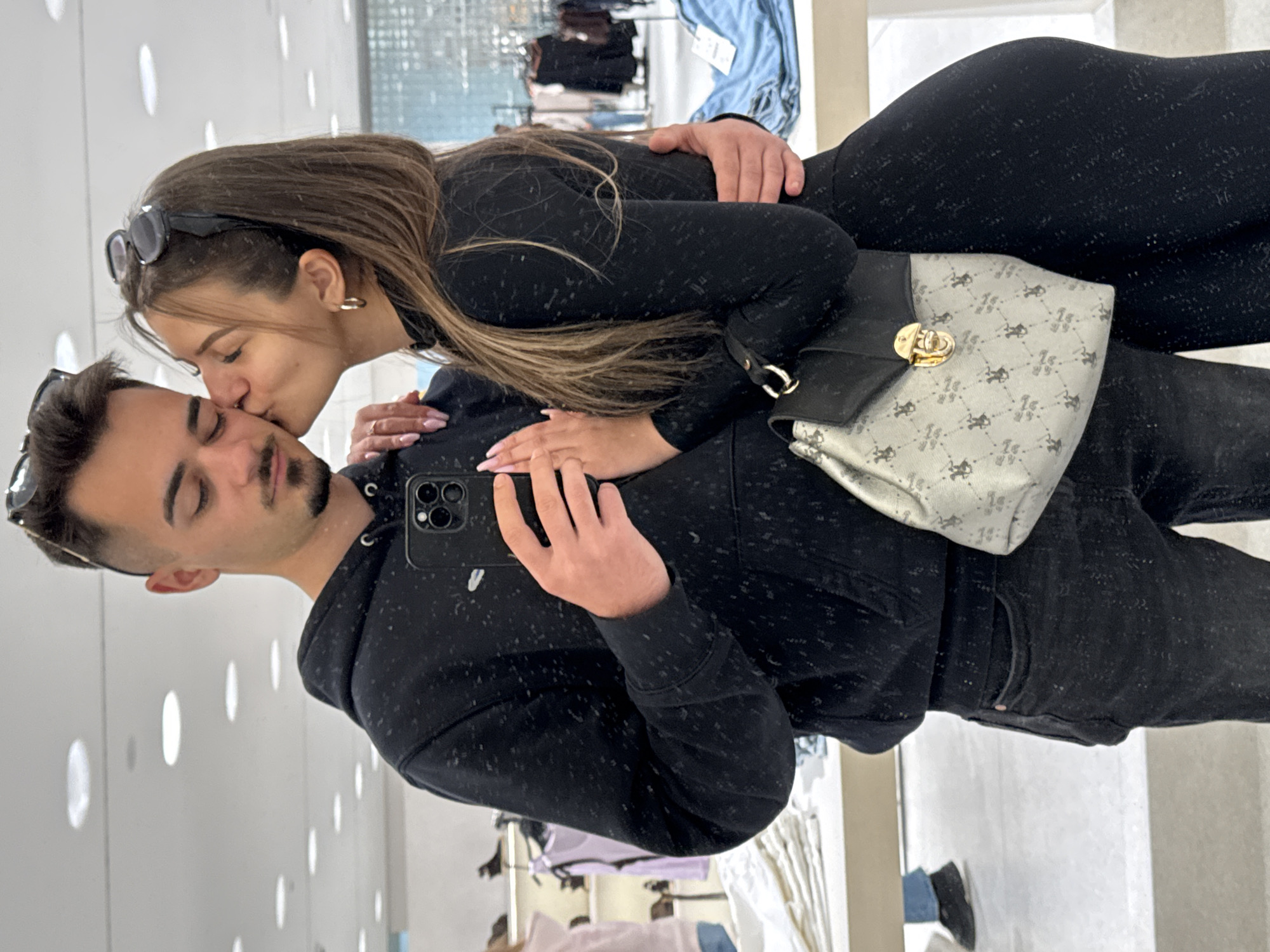 A woman kisses a man's head while he takes a selfie in a store. The man wears a black hoodie and black jeans, holding a black smartphone. The woman wears a black long-sleeved top and leggings. A patterned handbag sits on top of the man's hoodie. The store's interior features clothes racks in the background and circular lighting fixtures on the wall. Without additional information, the specific location in Sofia, Bulgaria, cannot be determined.