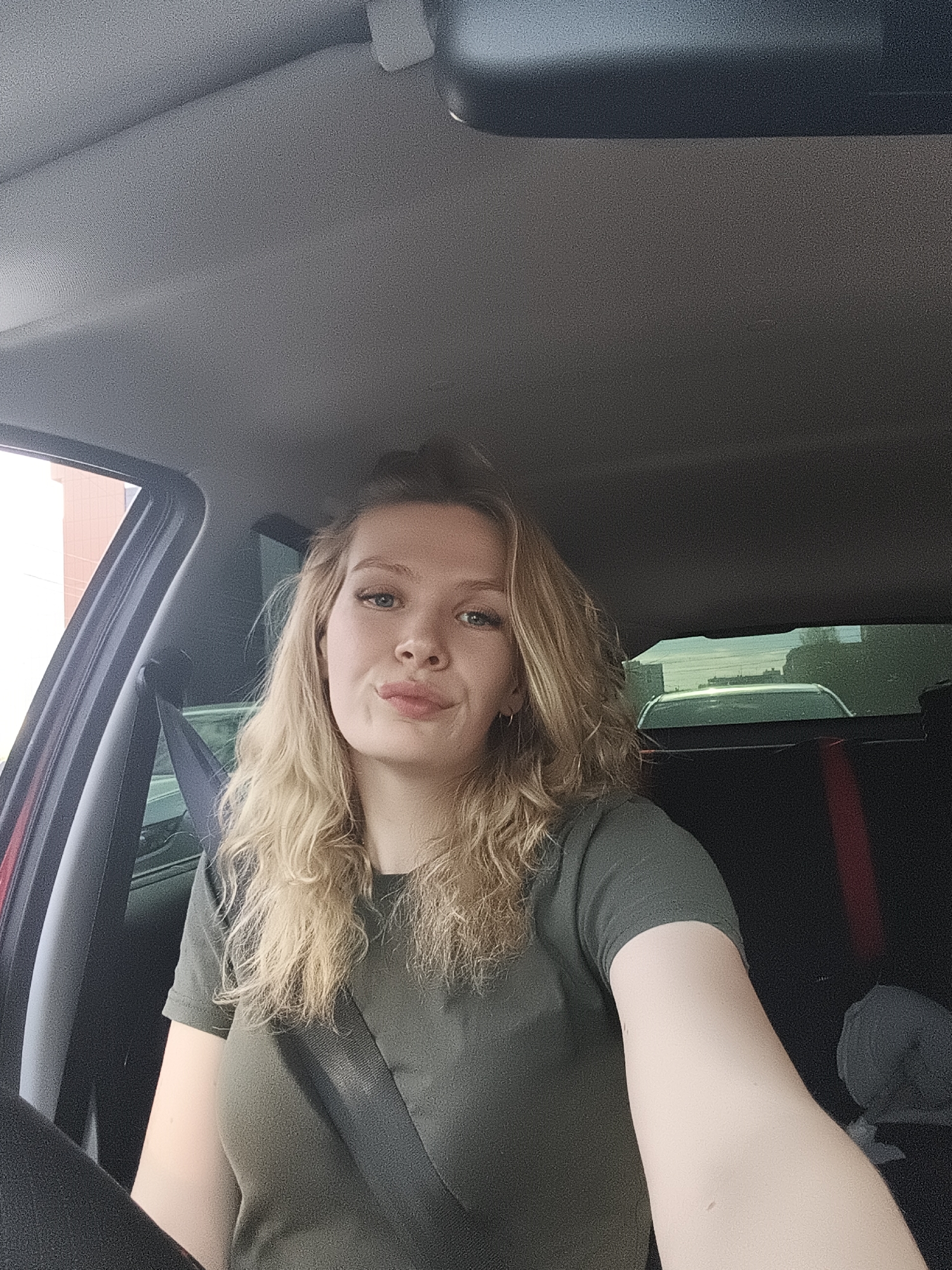 A young woman with blonde, wavy hair is sitting in the driver's seat of a car. She is wearing an olive green t-shirt and a seatbelt. She has fair skin and light blue eyes, and her lips are slightly pursed. The car's interior is visible, including the dashboard, steering wheel, and rearview mirror. The car is parked. The location in Izhevsk, Russia is not discernible from the image.